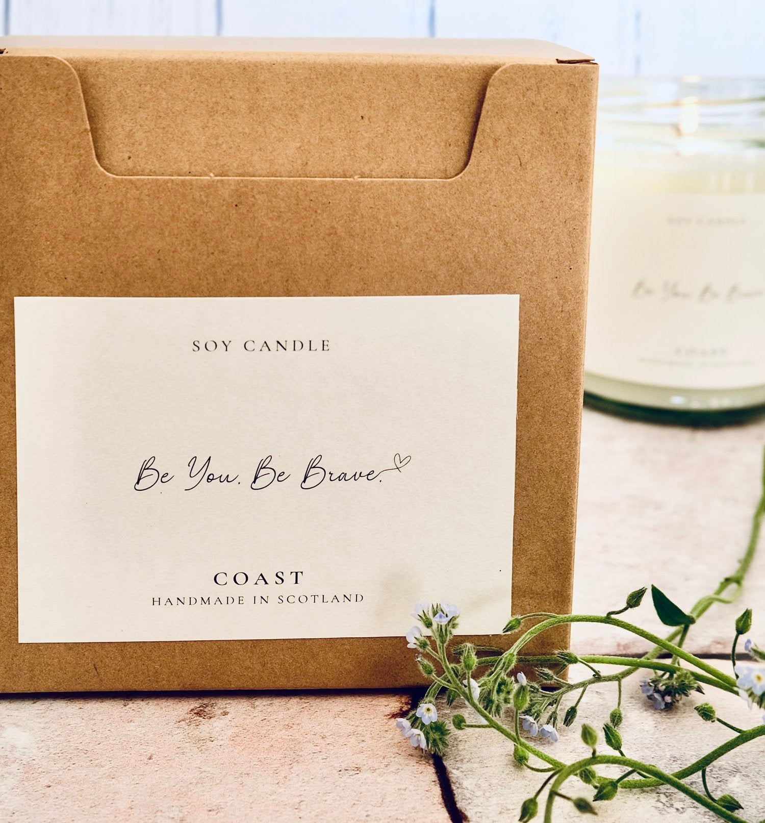 A gold lidded glass jar candle with white label sits in front of cardboard gift boxes with JoJo Co. candle branding. The main label reads 'Be you. Be brave'.