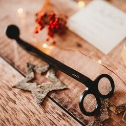 Matt black cast iron wick trimmers with a small JoJo Co. logo etched on them sits beside a wooden star on a pine floor