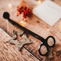 Matt black cast iron wick trimmers with a small JoJo Co. logo etched on them sits beside a wooden star on a pine floor
