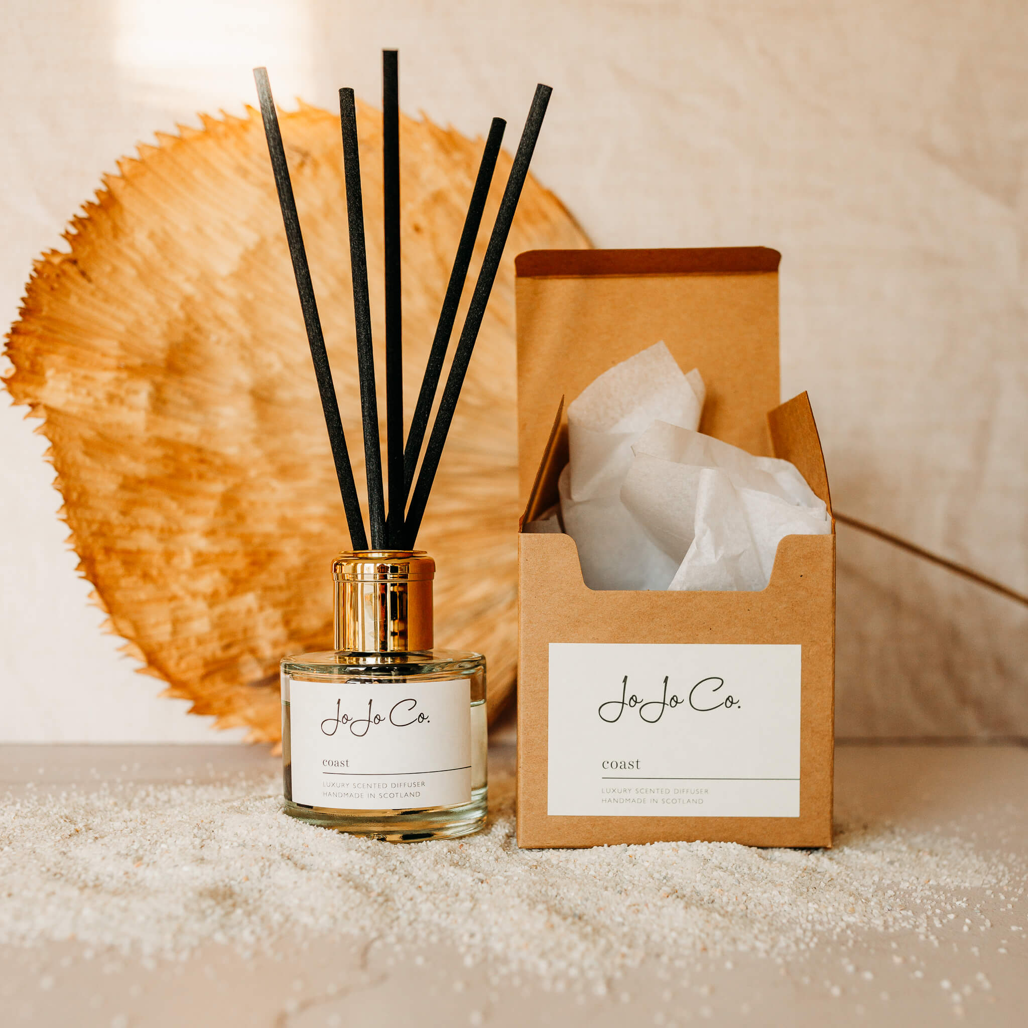 A glass diffuser with gold lid and black reeds sits on top of scattered sand next to a small open box and large dried leaf