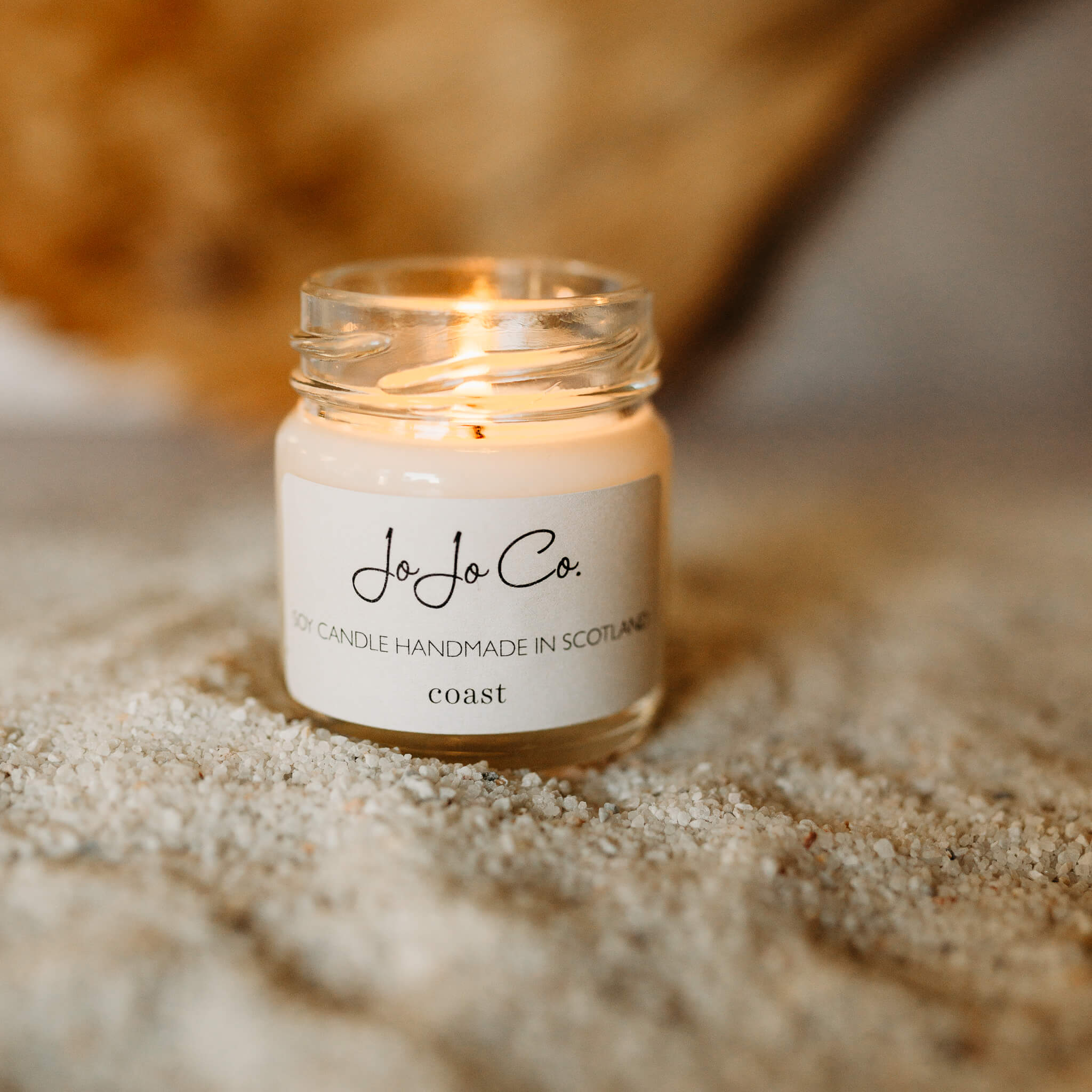 JoJo Co. mini Coast candle in a small glass jar and white label. The jar sits on top of sand, the detail of leaves in the background.