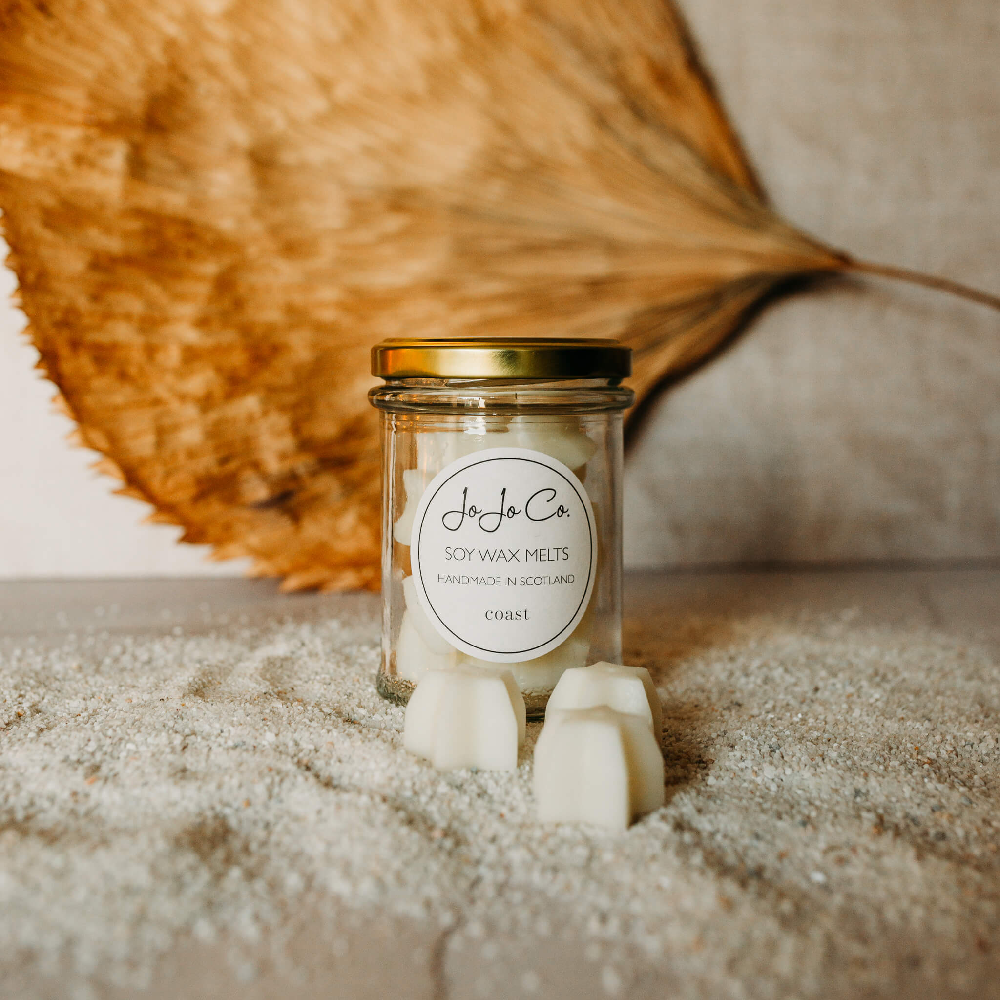 A glass jar with round JoJo Co. label filled with star shaped wax melts. In the foreground are three pretty wax star melts and a scattering of sand.