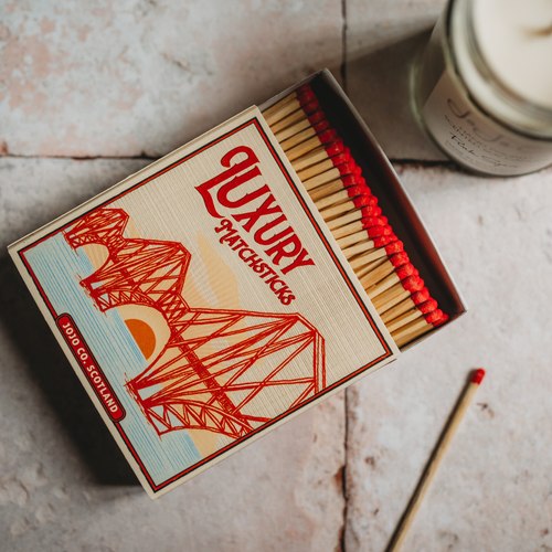 A large, square box of matches sits on cream tiles. Red matches are poking out and on the cover is a picture of the Forth Rail Bridge.