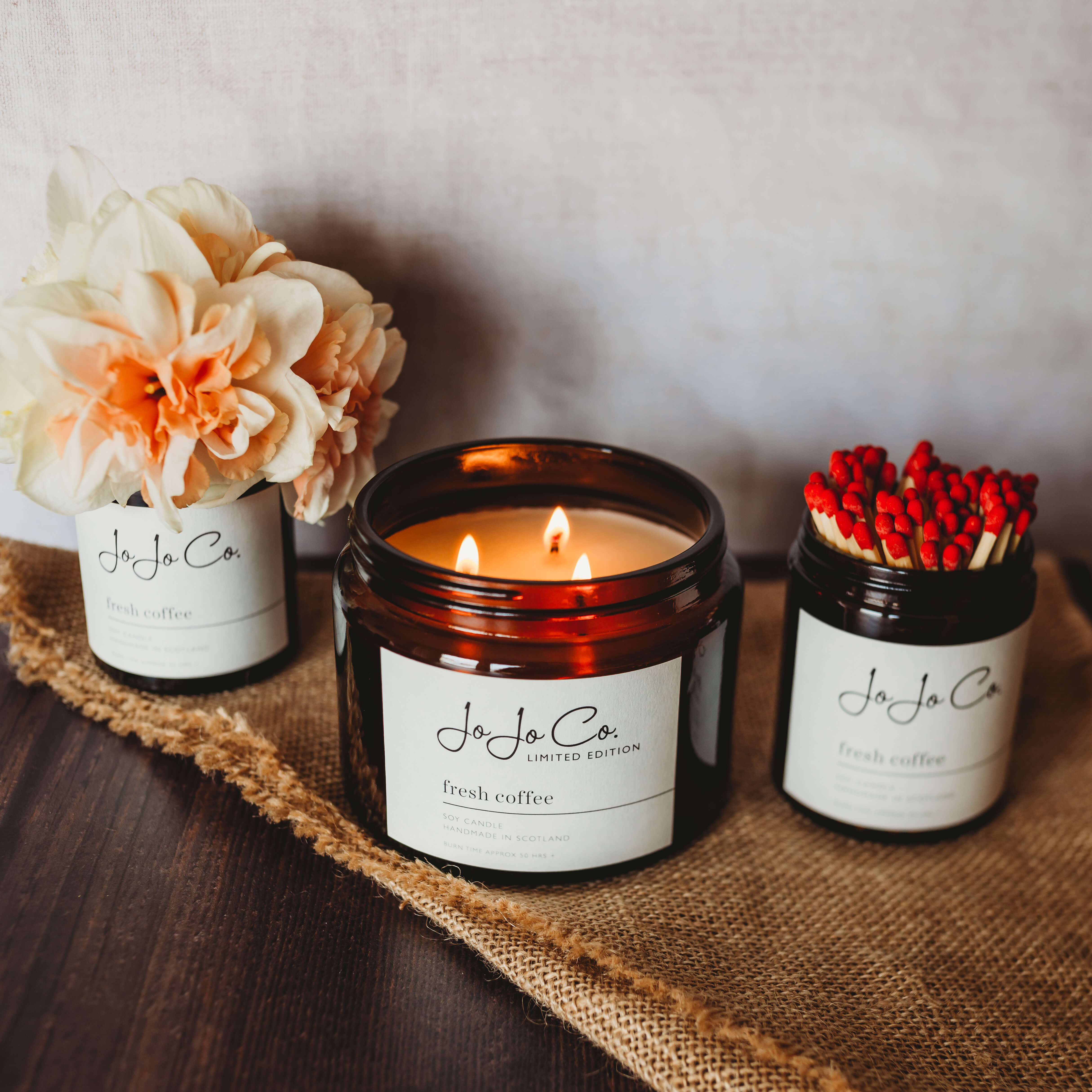 A small scene of amber jars. One contains long matchsticks with red heads. Another contains three-wick candle and flickering flames. The third contains a large, peach coloured flower.