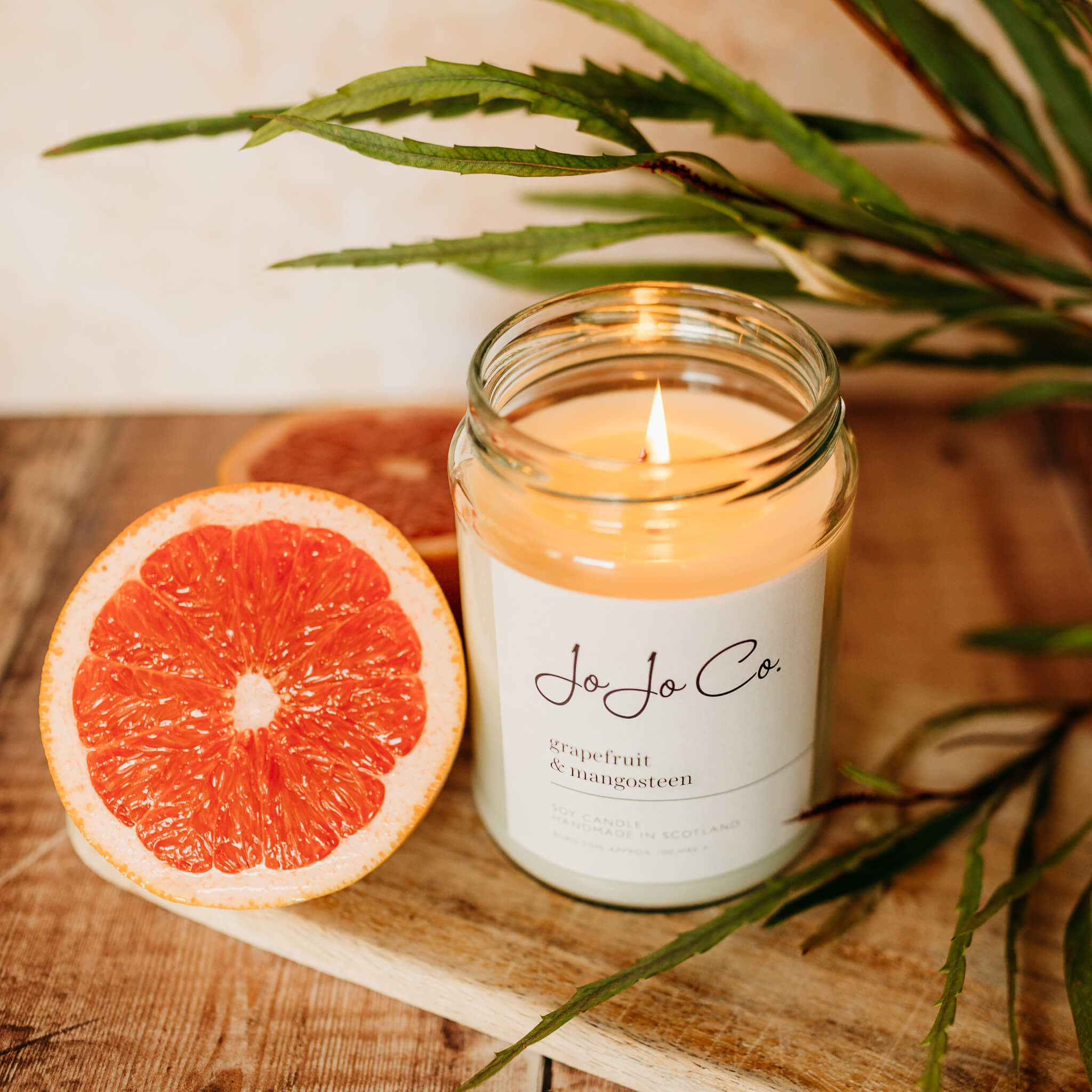 A brightly lit JoJo Co. Grapefruit & Mangosteen candle sits on a wooden surface among slices of pink grapefruit and big green leaves.