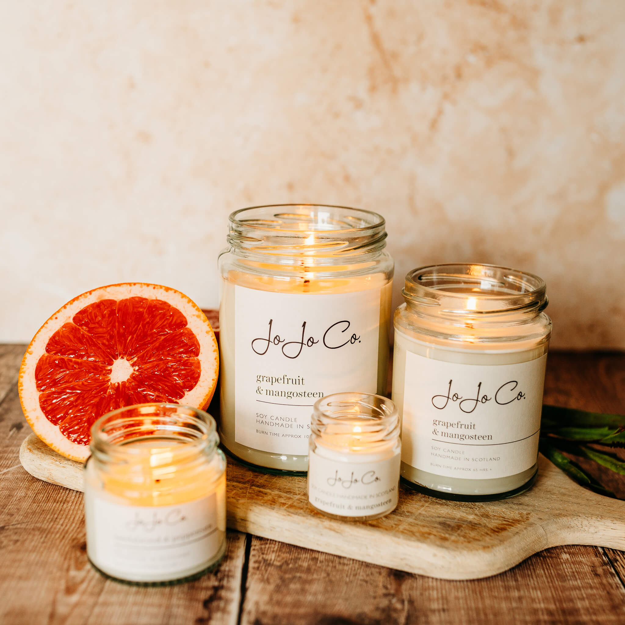 A selection of four JoJo Co Grapefruit & Mangosteen candles sit on a wooden chopping board with peach marbled background. A slice of juicy grapefruit sits beside them.
