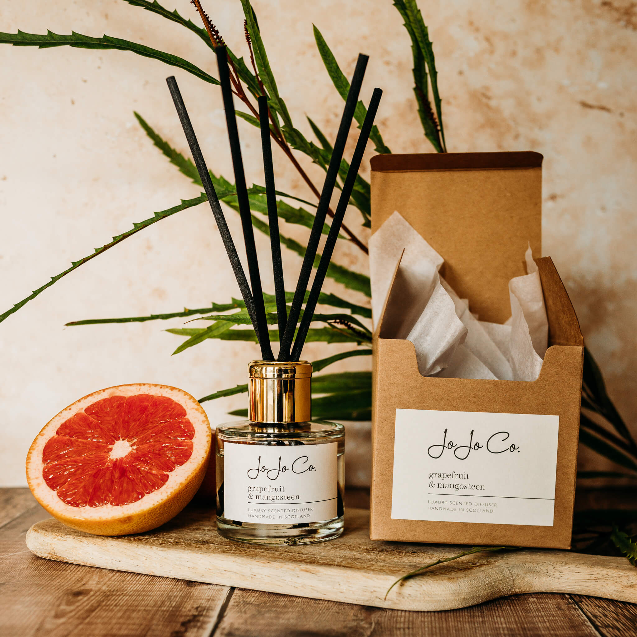 A glass diffuser with JoJo Co. white label, gold lid and black reeds sits on a wooden chopping board in front of a lush green plant. Beside the diffuser is a slice of ruby grapefruit and an open gift box with white tissue paper inside.