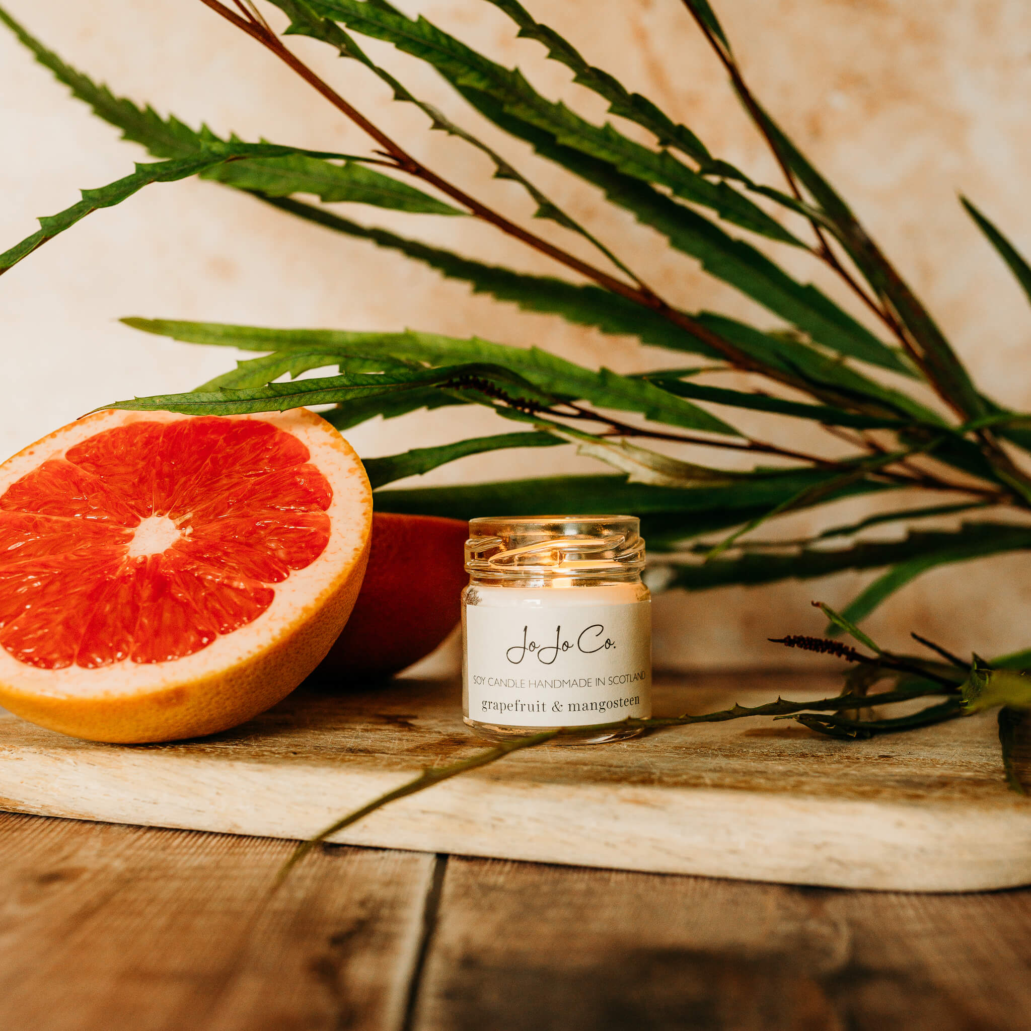 A mini glass jar JoJo Co. Grapefruit & Mangosteen candle sits on a wooden chopping board with leaves and grapefruit surrounding it.