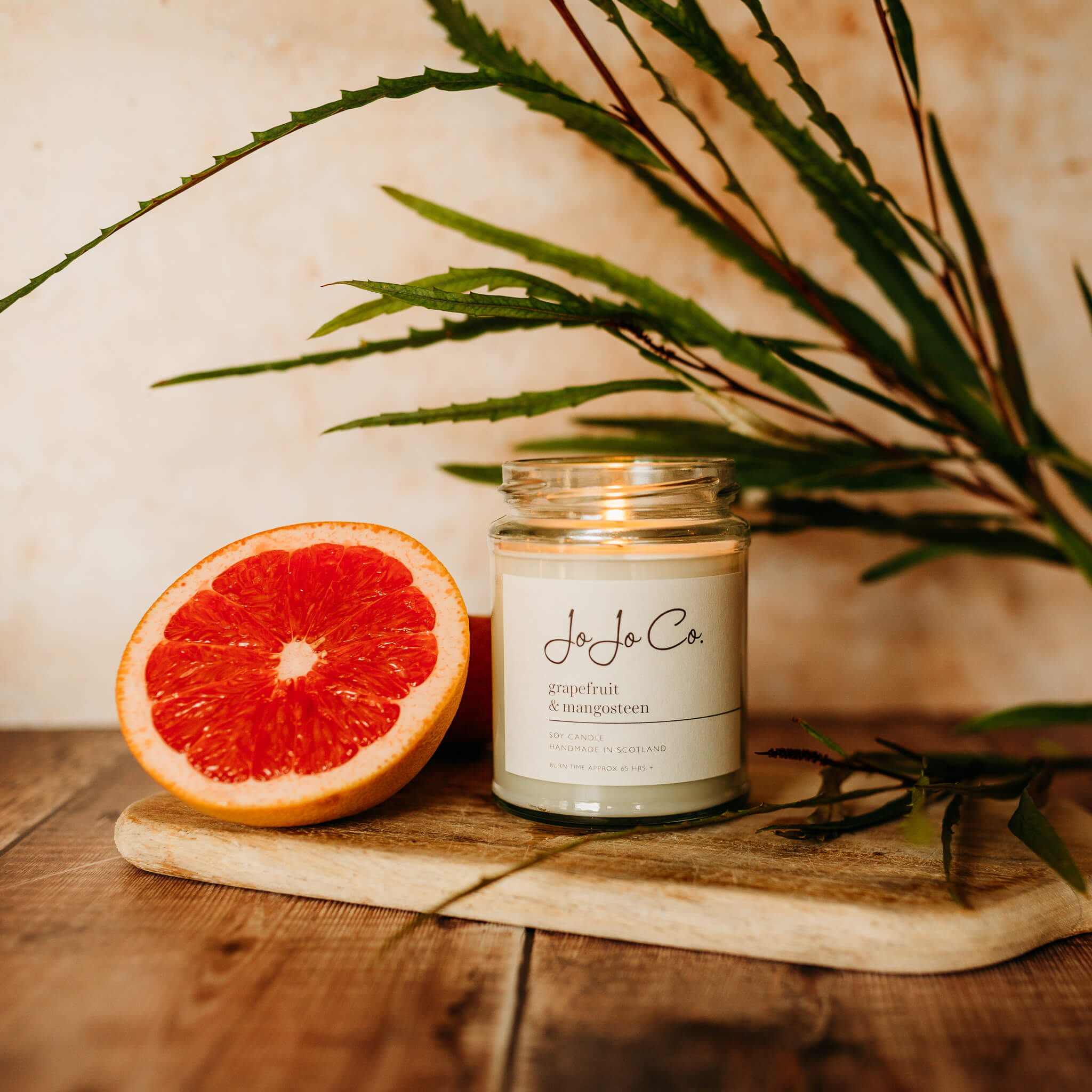 A large glass JoJo Co Grapefruit & Mangosteen candle sits beside a slice of pink grapefruit on a wooden chopping board. A large green leaf leans against a peach stone background