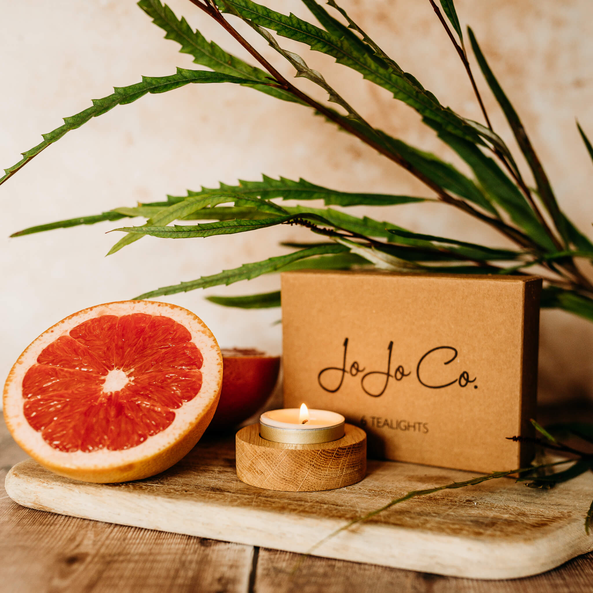 A lit tealight sits in a round oak tealight base. In the background is a cardboard gift box with JoJo Co. logo printed on top. A green plant and slices of ruby grapefruit sit beside it.