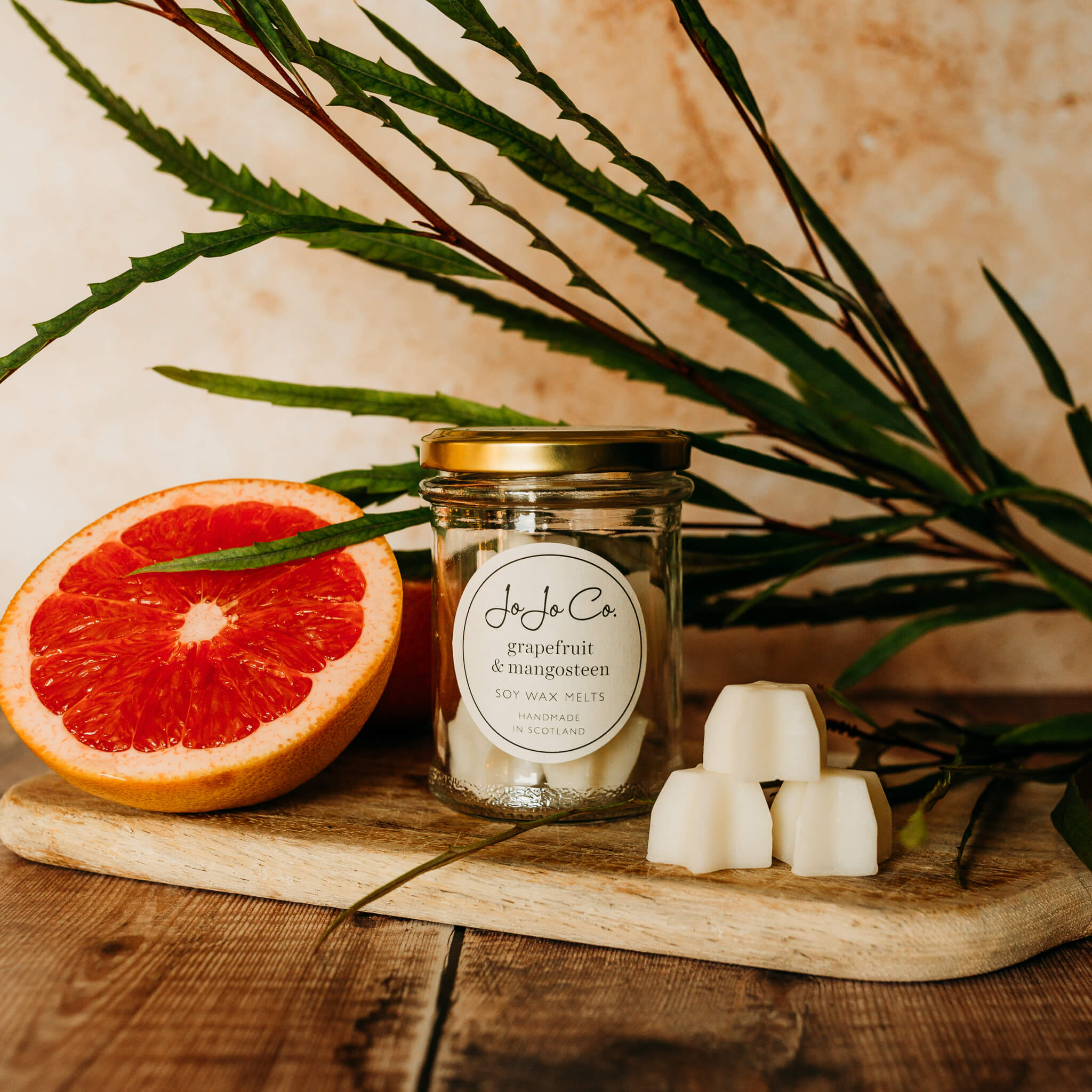 A glass jar with gold lid and white JoJo Co. label sits on top of a wooden chopping board in front of a green plant. Beside the jar is a slice of ruby grapefruit and a trio of star shaped wax melts.