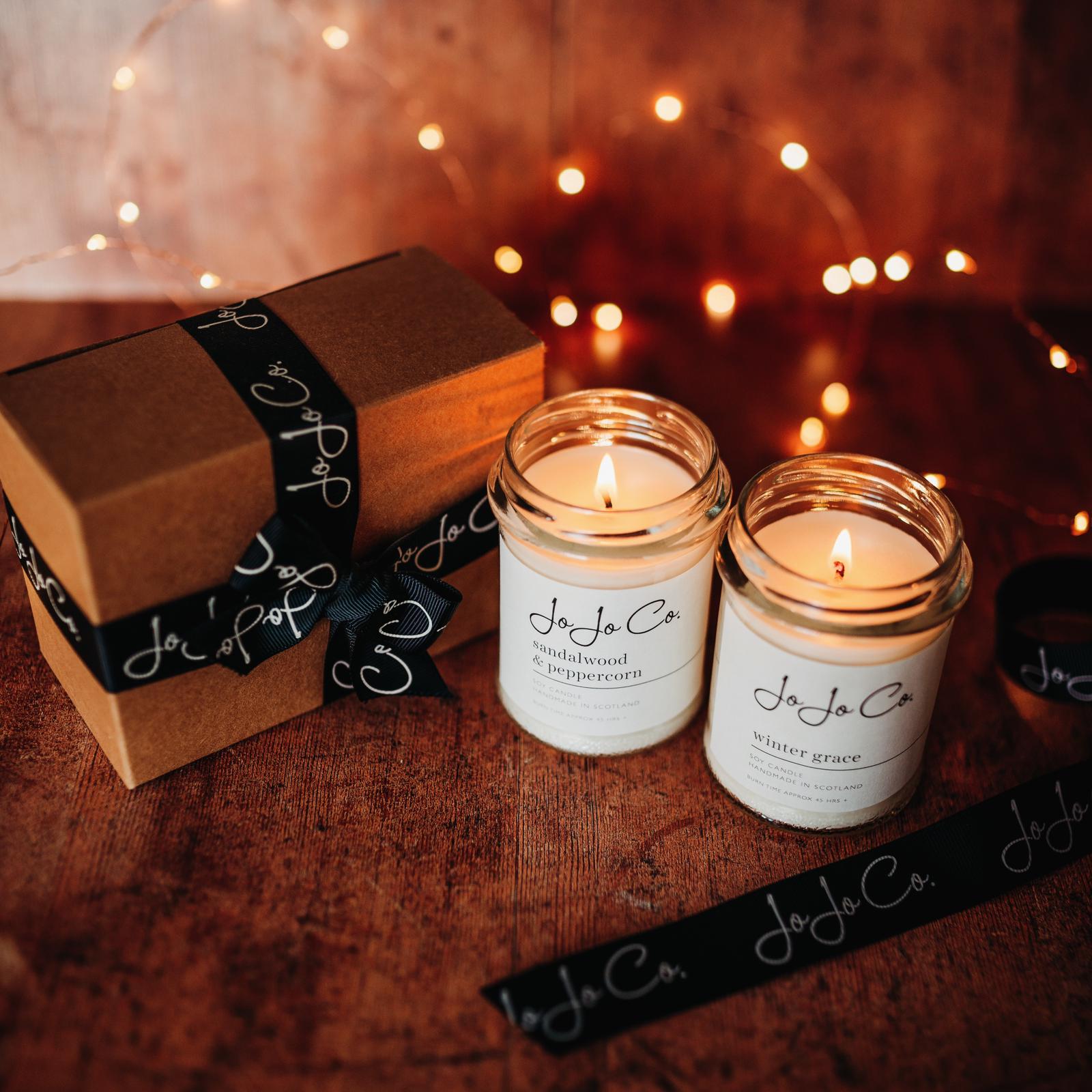 Two glass candles with white labels and JoJo Co logo sit beside a cardboard box with black bow in front of warm fairy lights in a warm and welcoming scene.