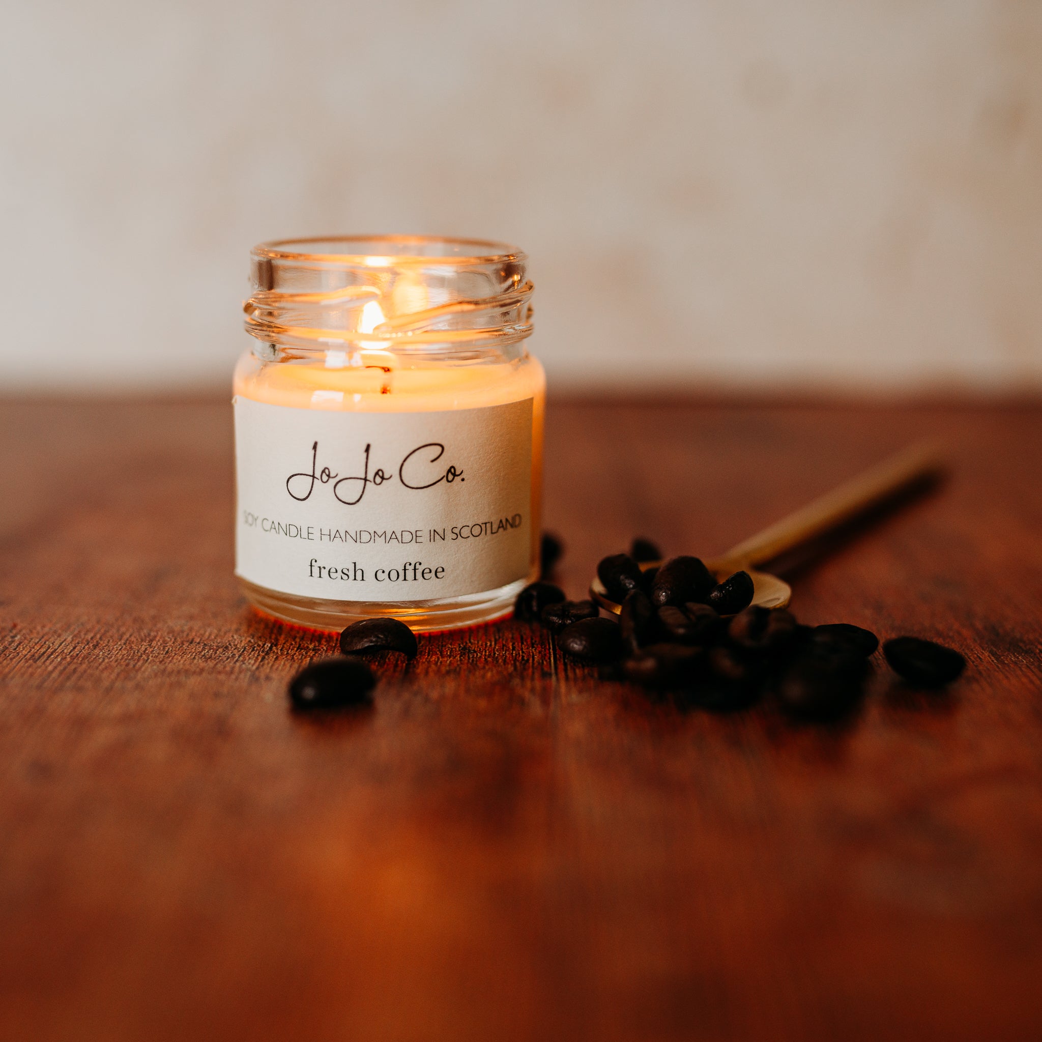 A small glass jar candle flickers with a warm glow from the flame. Beside it sits a golden spoon and a scatter of coffee beans.