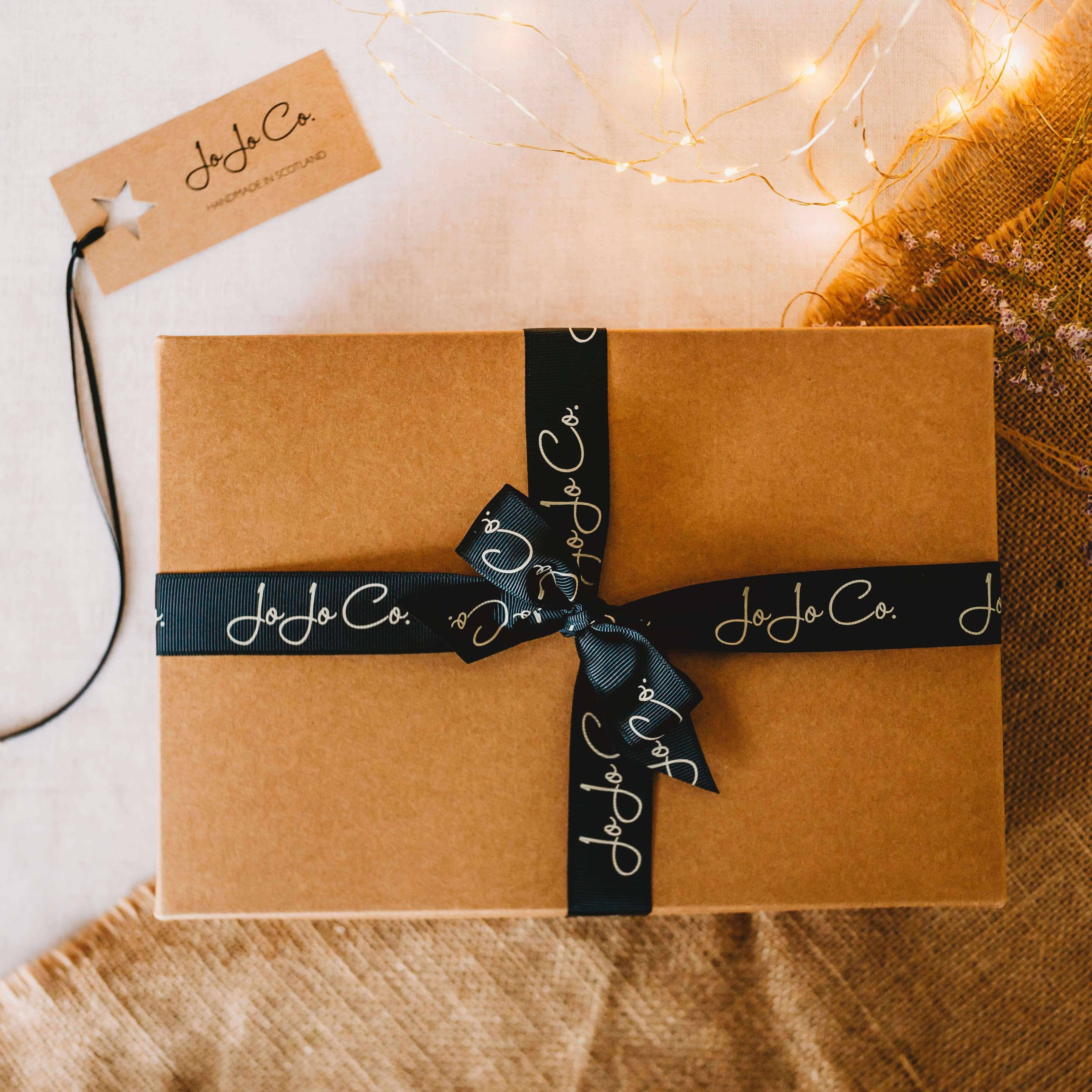 A cardboard gift box tied with black JoJo Co. bow sits on top of jute fabric, with fairy lights and gift tag in the image.