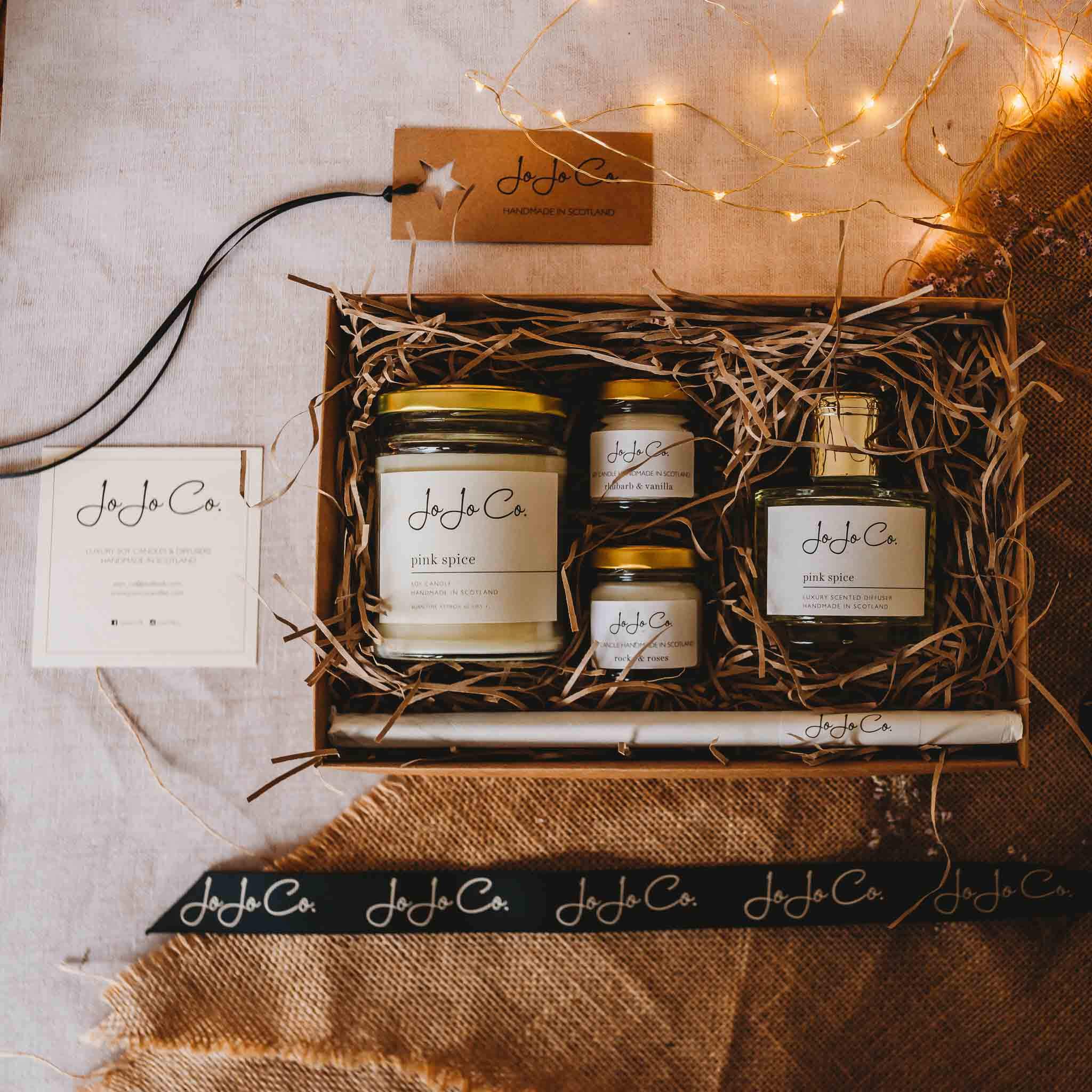 A selection of candle and diffuser products from JoJo Co. Three glass jar candles with gold lids, a glass jar diffuser with gold lid, wrapped reeds. The box is surrounded by fairy lights, JoJo Co. ribbons, a gift tag and jute fabric.