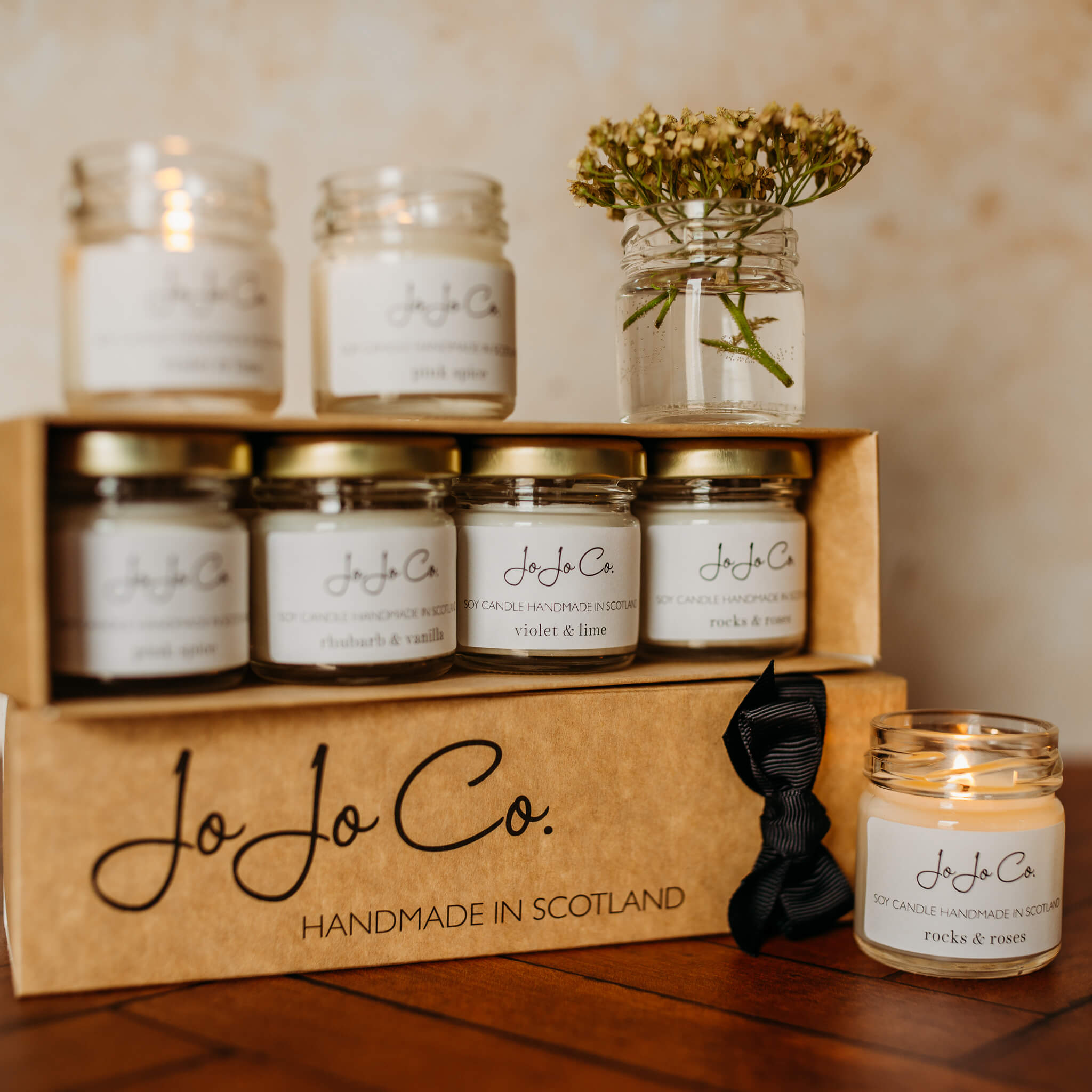 A neat cardboard gift box containing four mini candles with gold lids. Below the box is another with a black bow, and two mini candles n the top next to a small pot of flowers.