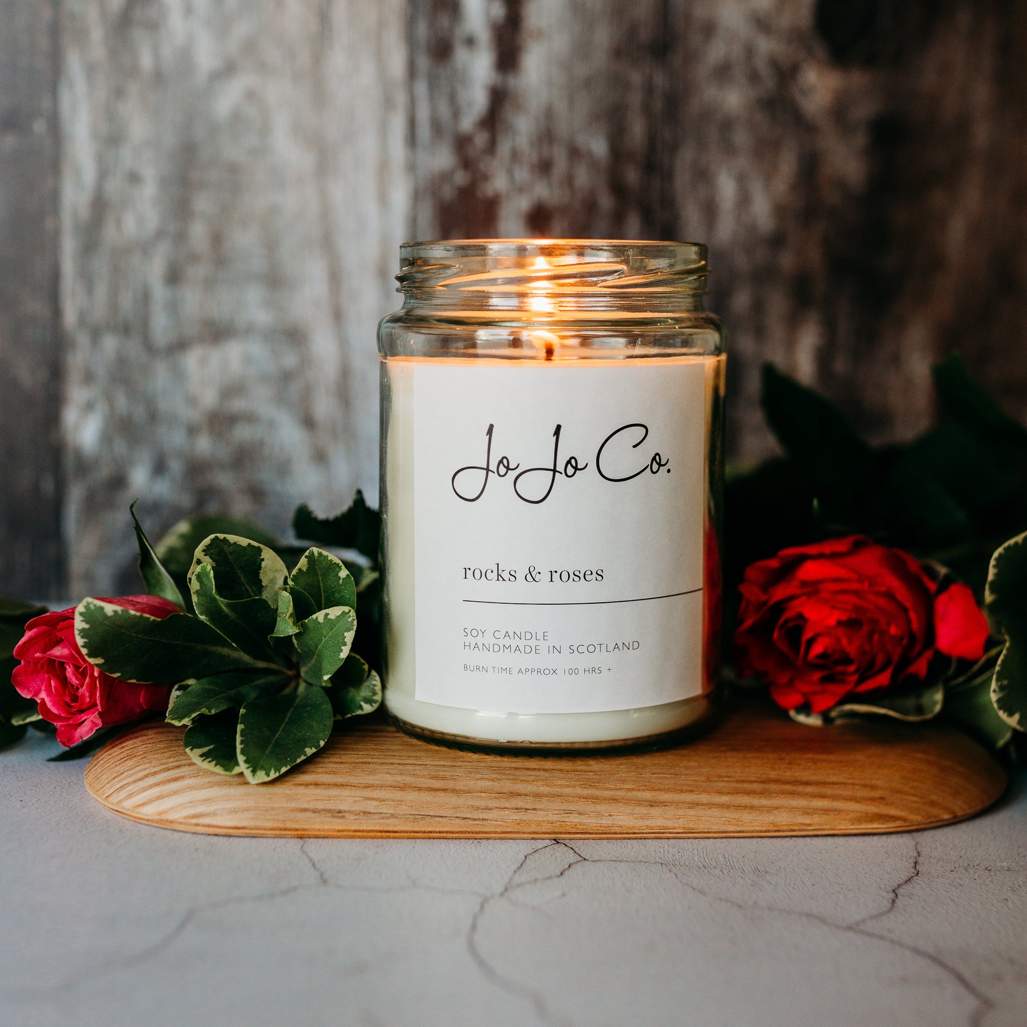 A close-up of a 100 hour glass candle jar with the label "JoJoCo rocks & roses" on a wooden surface, with green leaves and red rose petals surrounding it, creating an intimate ambiance.