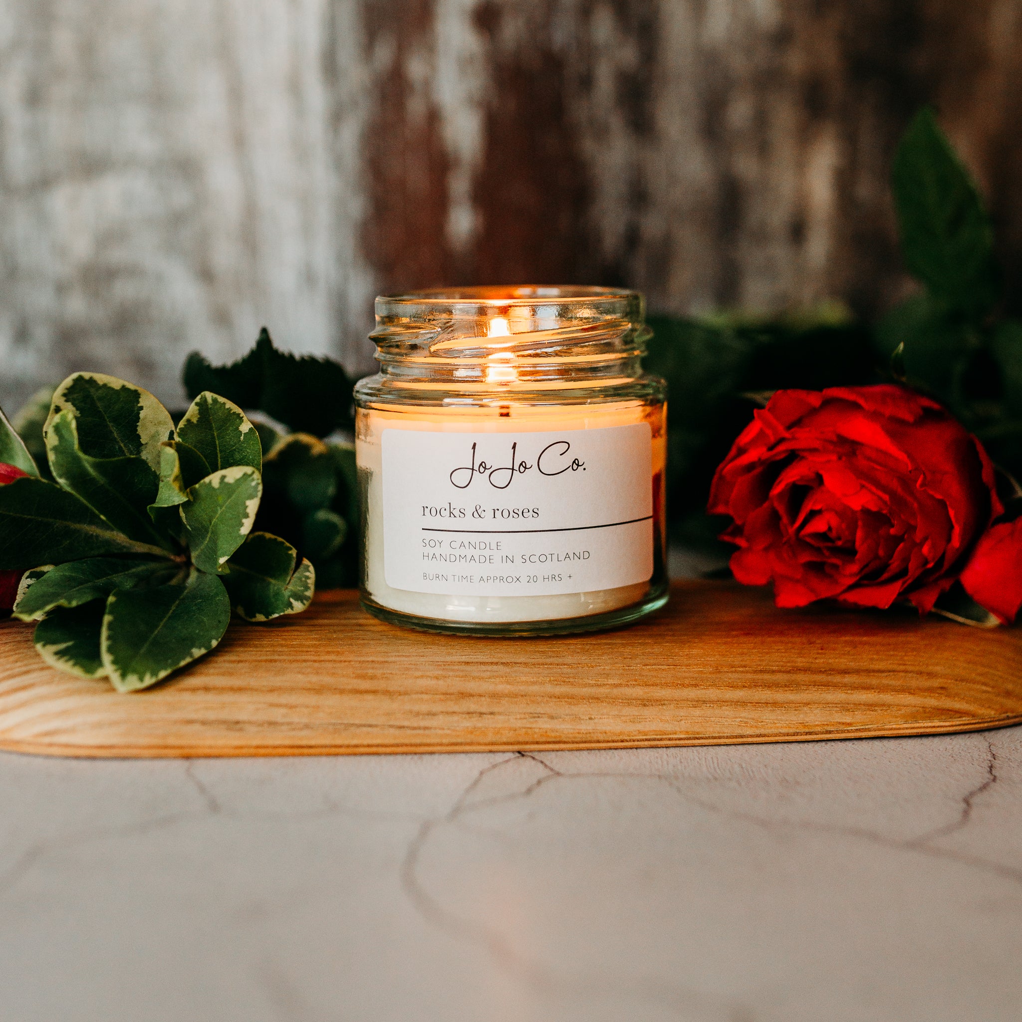 A close-up of a 20 hour glass candle jar with the label "JoJoCo rocks & roses" on a wooden surface, with green leaves and red rose petals surrounding it, creating an intimate ambiance.
