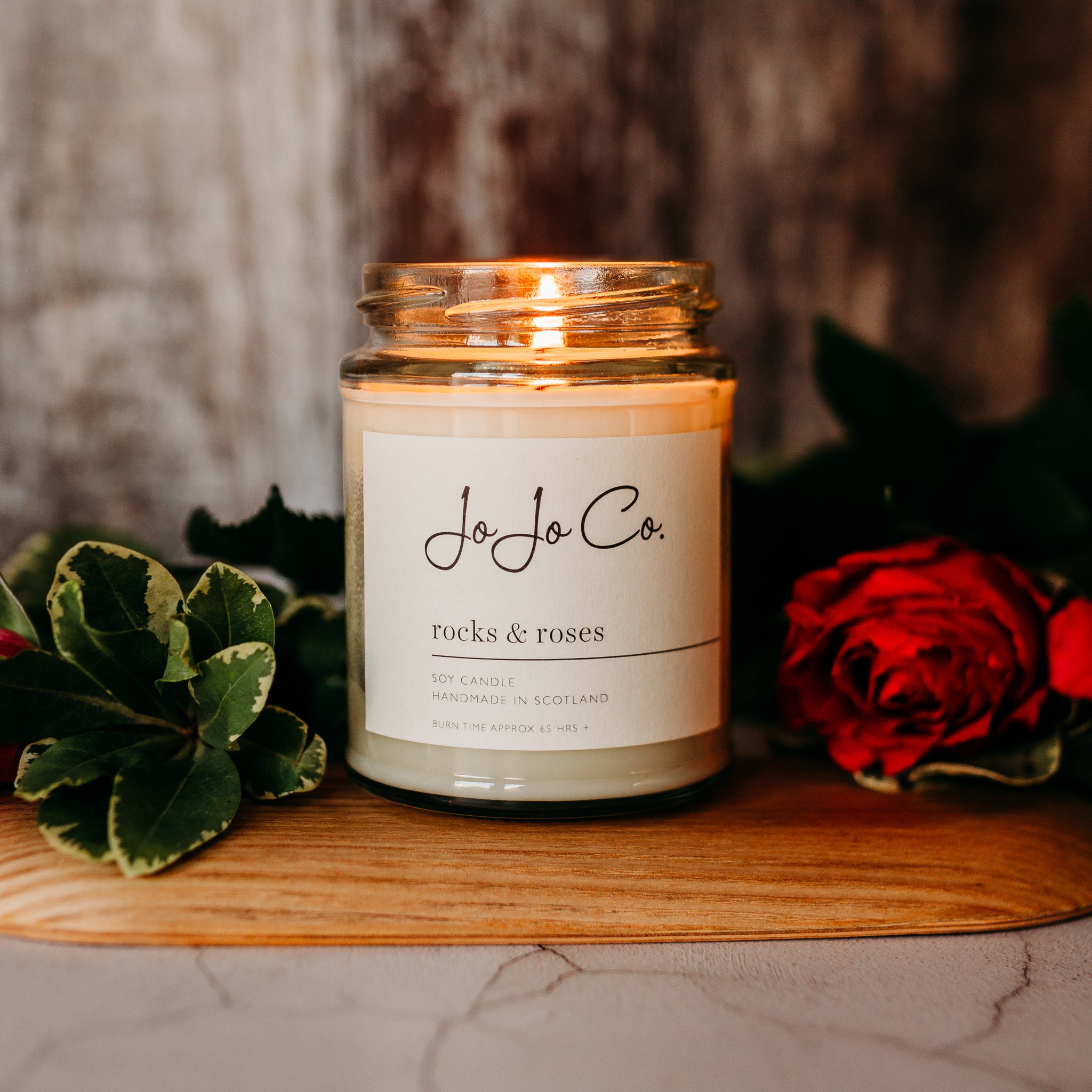 A close-up of a 65-hour glass candle jar with the label "JoJoCo rocks & roses" on a wooden surface, with green leaves and red rose petals surrounding it, creating an intimate ambiance.