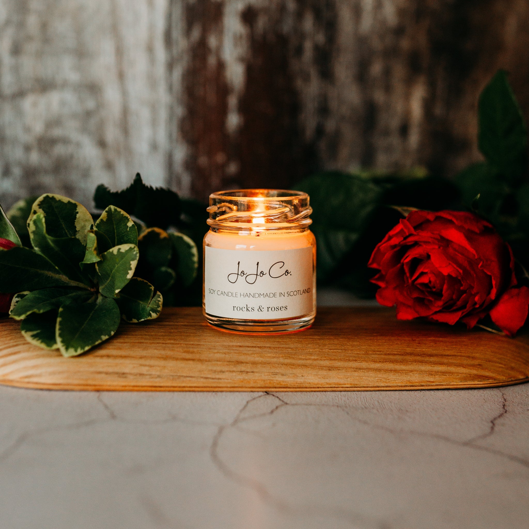 A close-up of a glass candle jar with the label "JoJoCo rocks & roses" on a wooden surface, with green leaves and red rose petals surrounding it, creating an intimate ambiance.