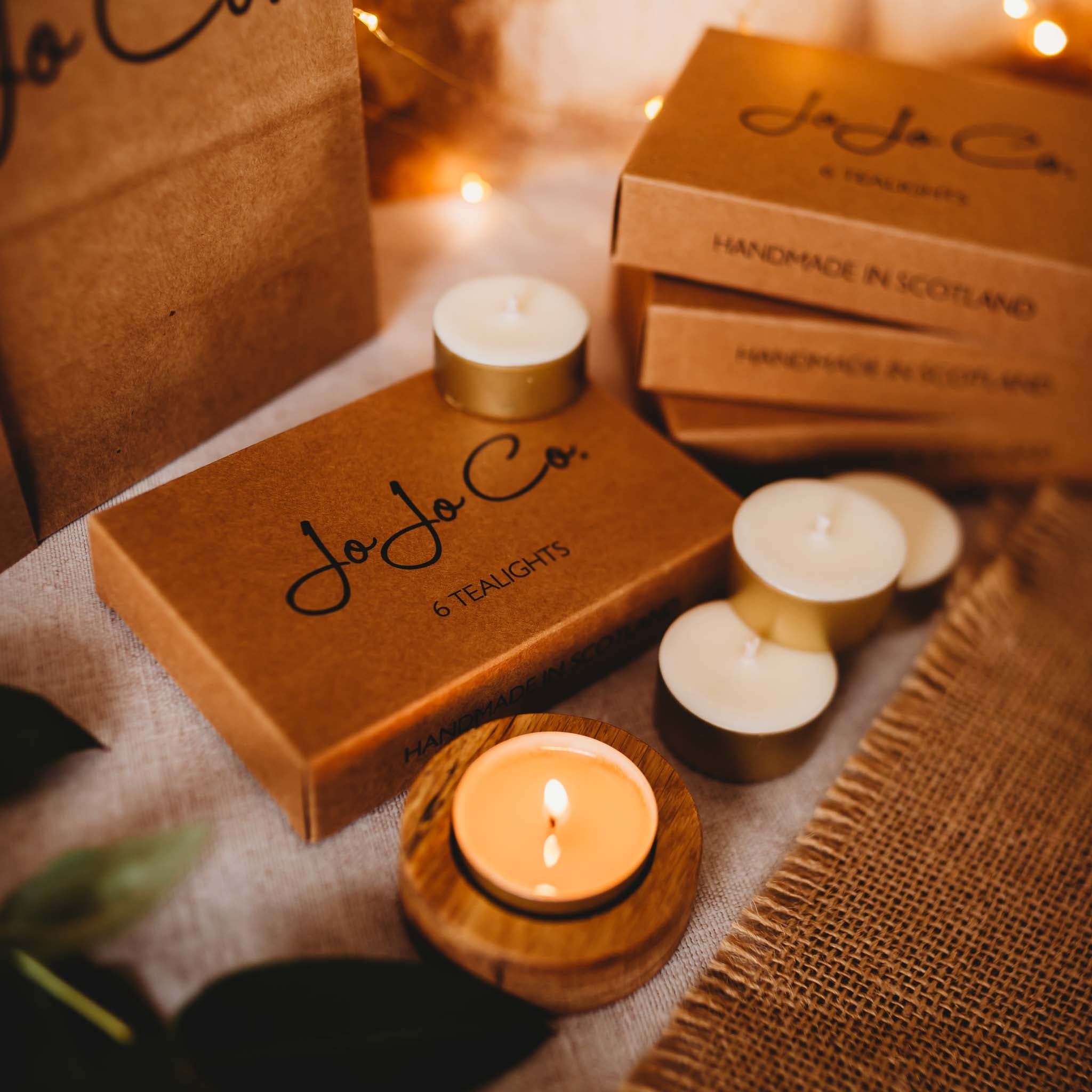 A lit tealight sits in a round oak tealight holder surrounded by unlit tealights, JoJo Co. branded gift boxes and fairy lights in the background