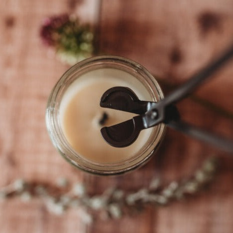 A close up of black, open wick trimmers over the top of a white candle as if to trim the wick