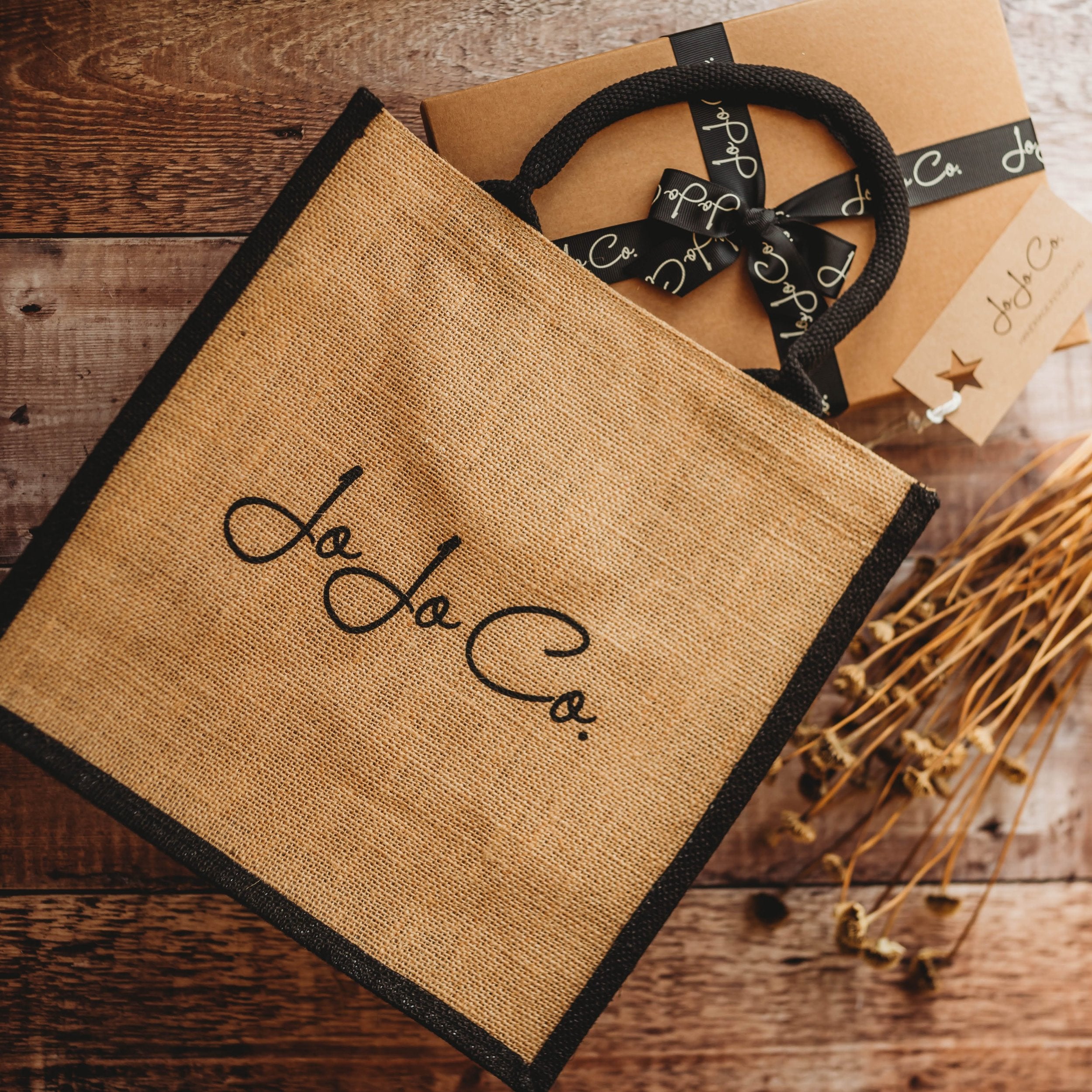 A natural coloured jute bag with black JoJo Co. logo and black trimming sits on top of a cardboard gift box with black branded bow.