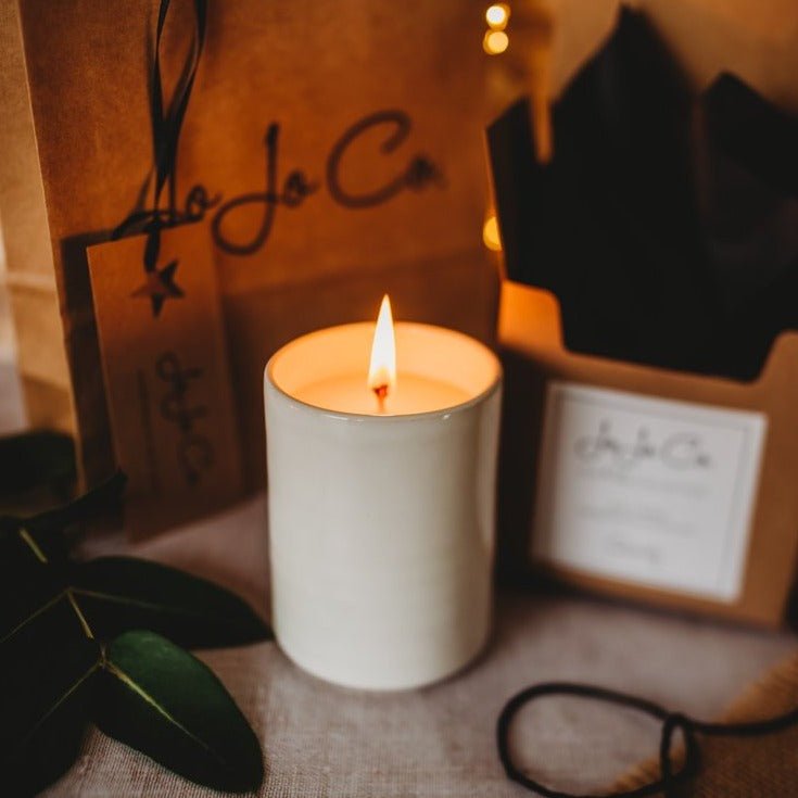 A lit candle inside a cream pottery jar. Beside the candle is a lush green leaf, various cardboard gift boxes with JoJo Co branding.