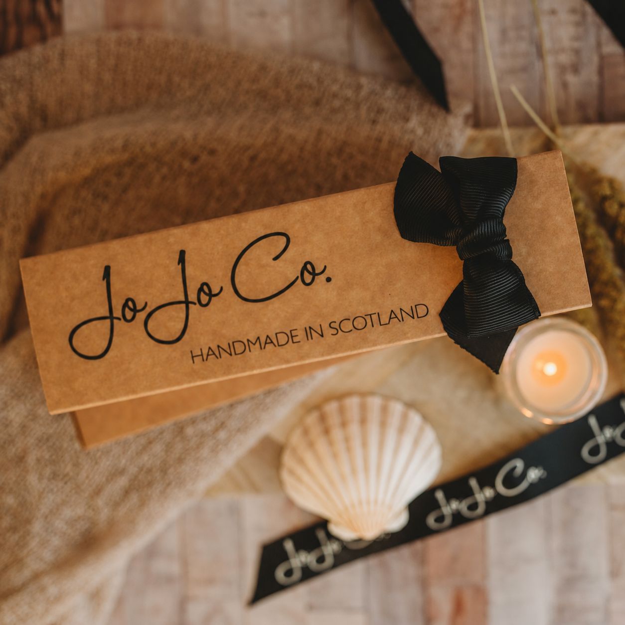 JoJo Co. packaging with a black ribbon, shell, and candle on a textured surface