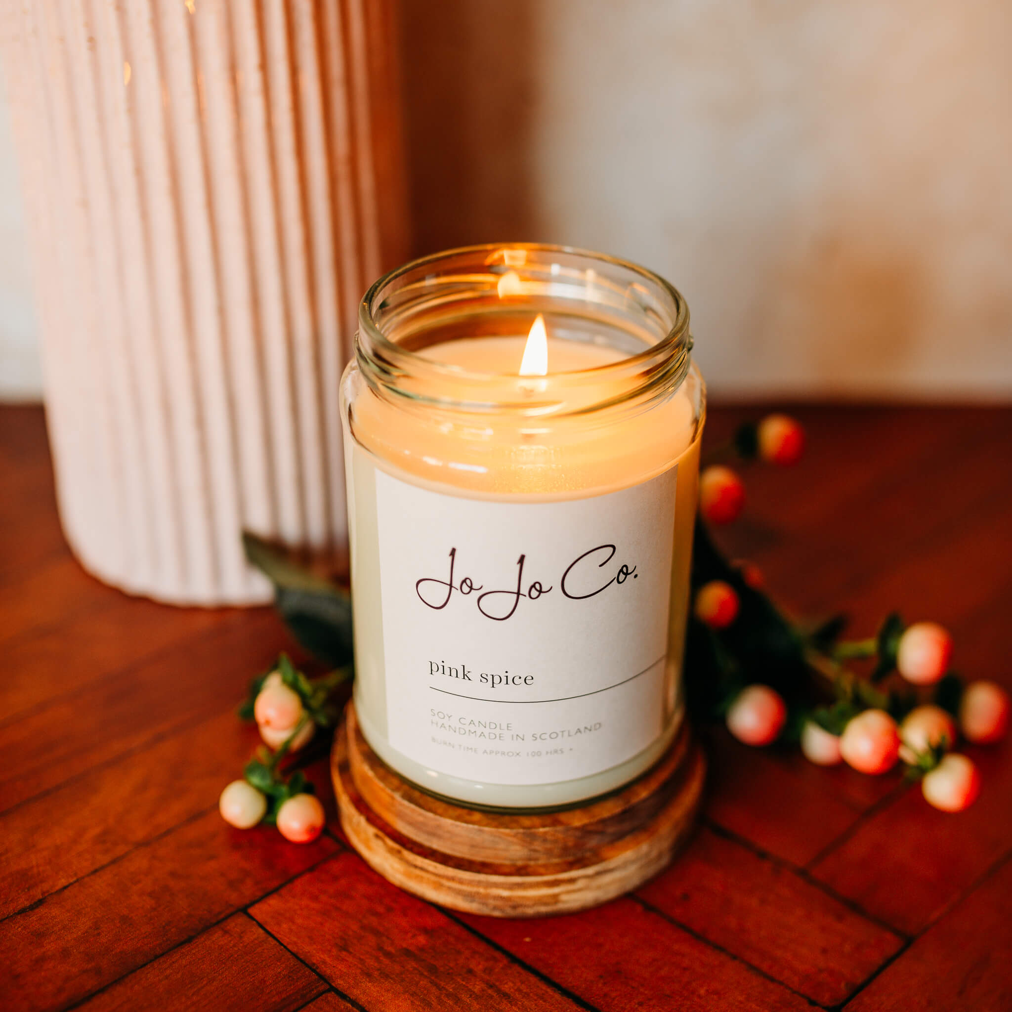 JoJo Co. Pink Spice 100 hour candle sits on wooden floor with pink berries in the background. The candle is lit