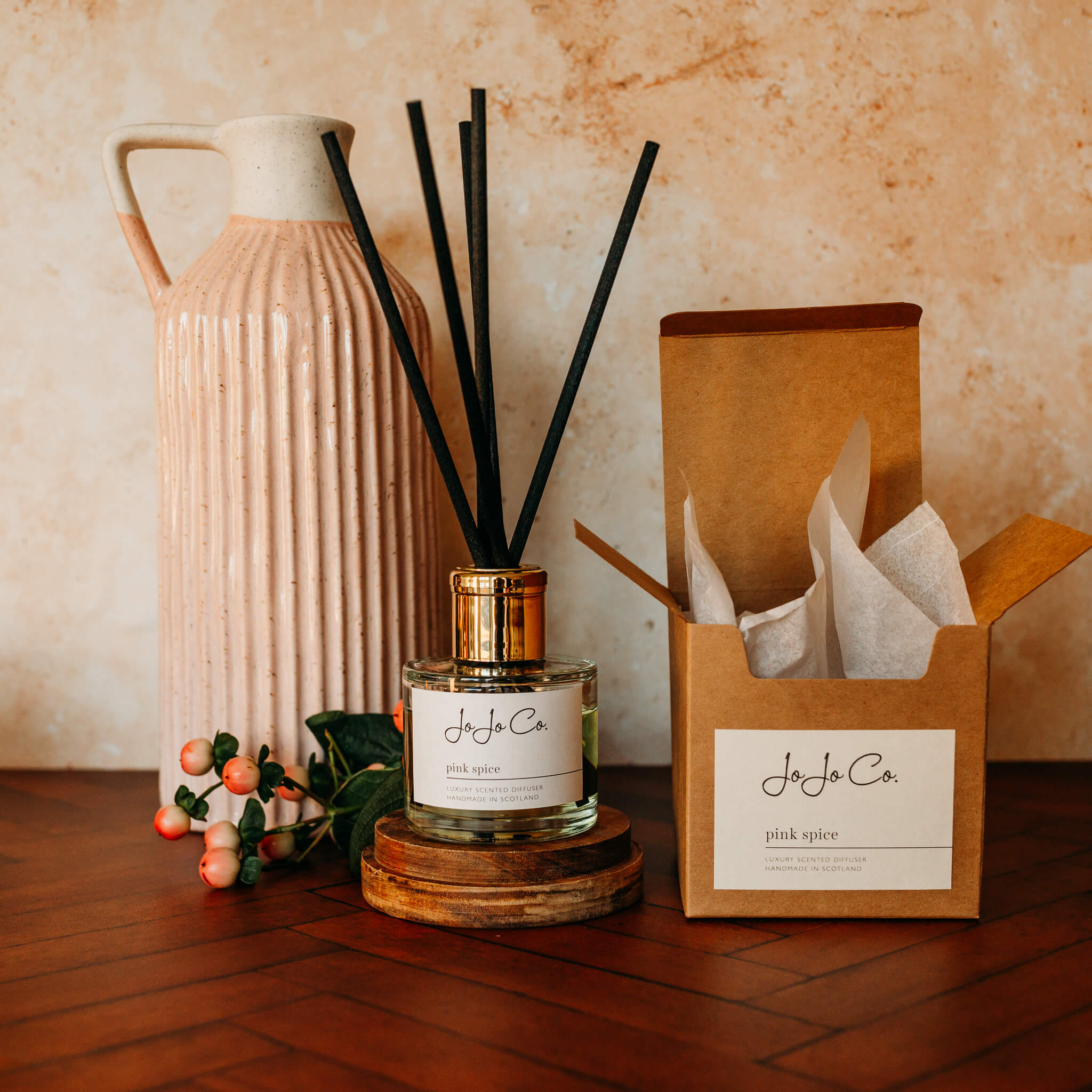 A glass jar diffuser with cream JoJo Co label, gold lid and black reeds sit on an oak candle base on a wooden floor. In the background is a pink and cream jug and pink blush berries on green stalks.