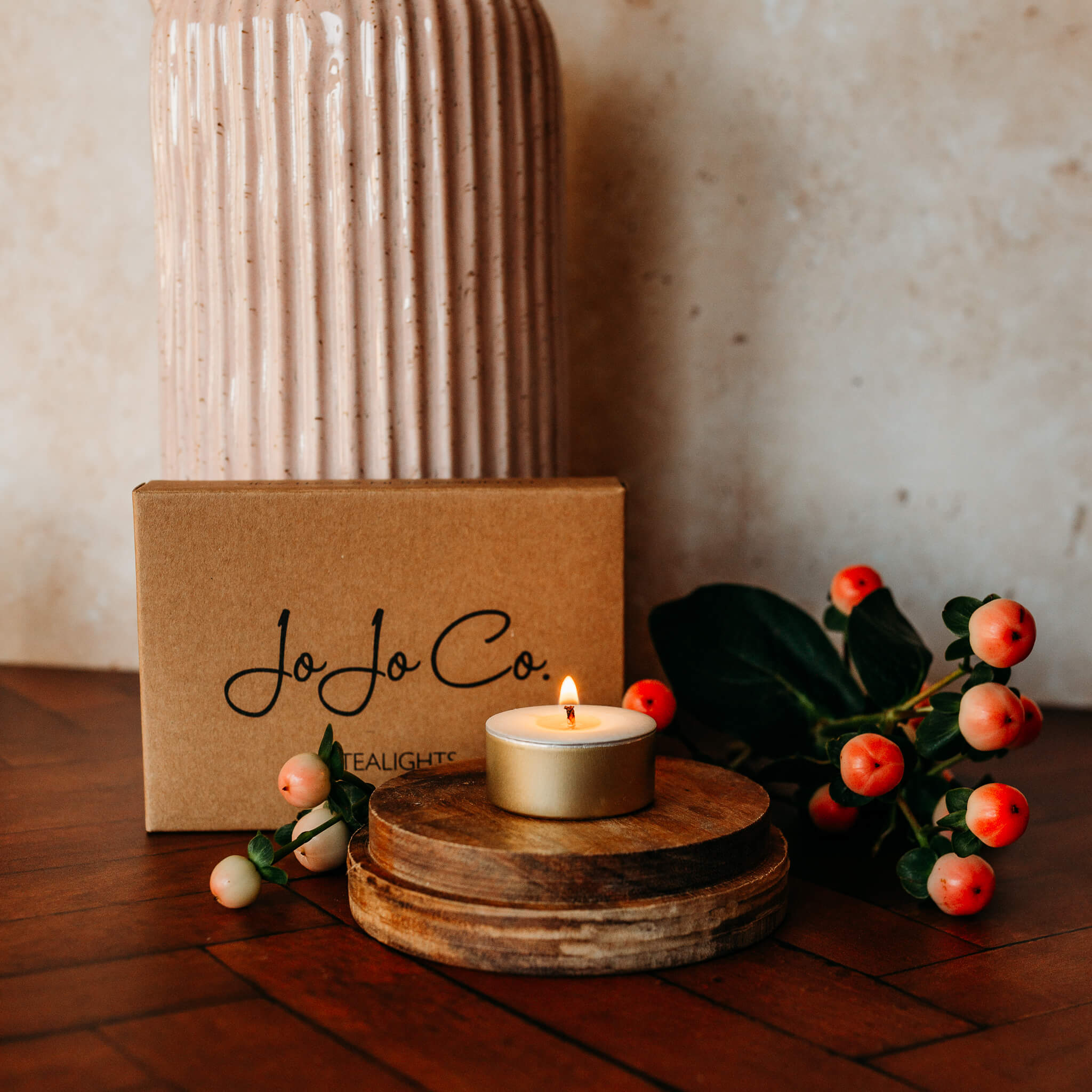 A lit tealight sits on a large oak base on a wooden floor. Behind the tealight is a cardboard gift box with JoJo Co logo, pink berries and a pink and cream jug.