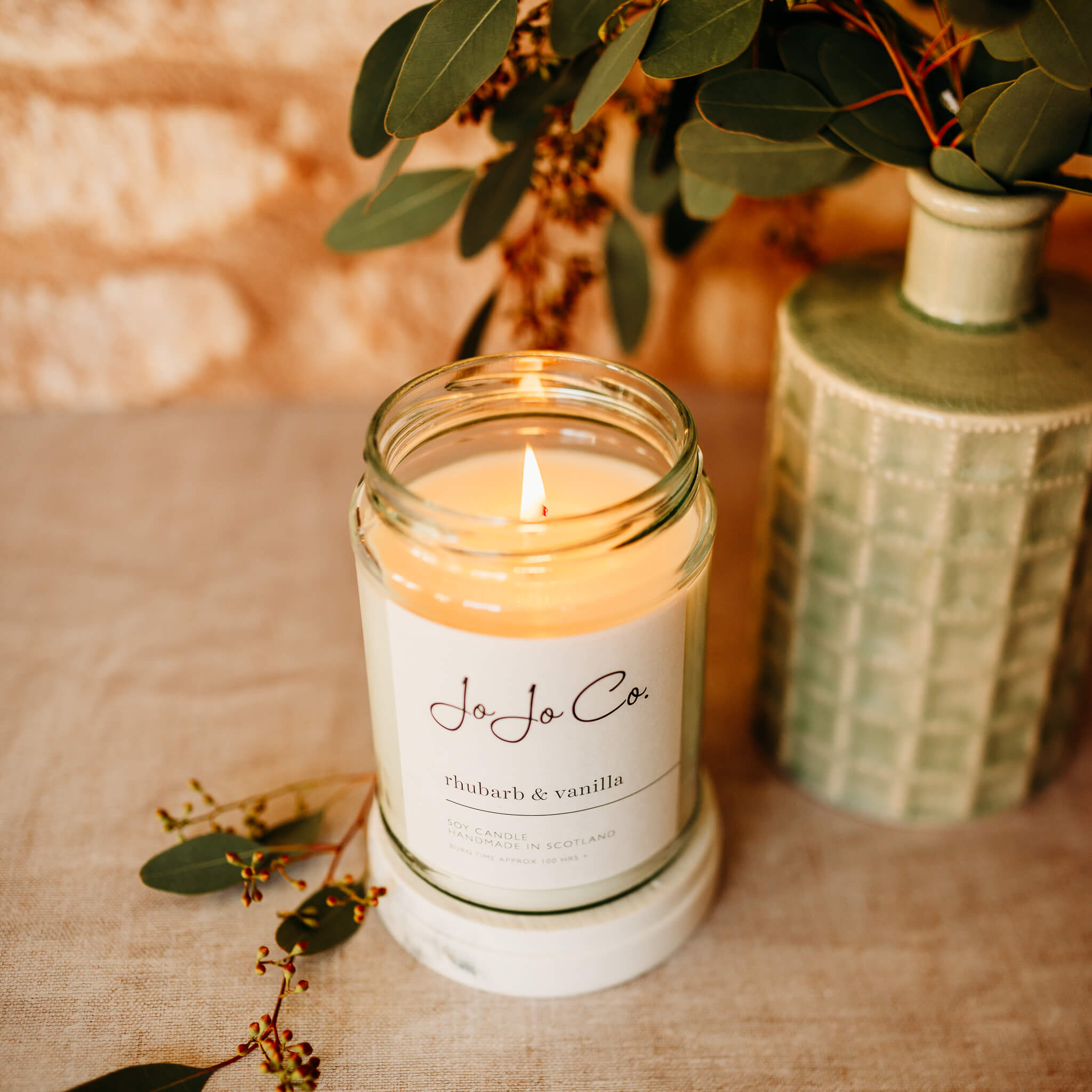 A lit JoJo Co 100 hour Rhubarb and Vanilla candle in a glass jar with a vase and foliage