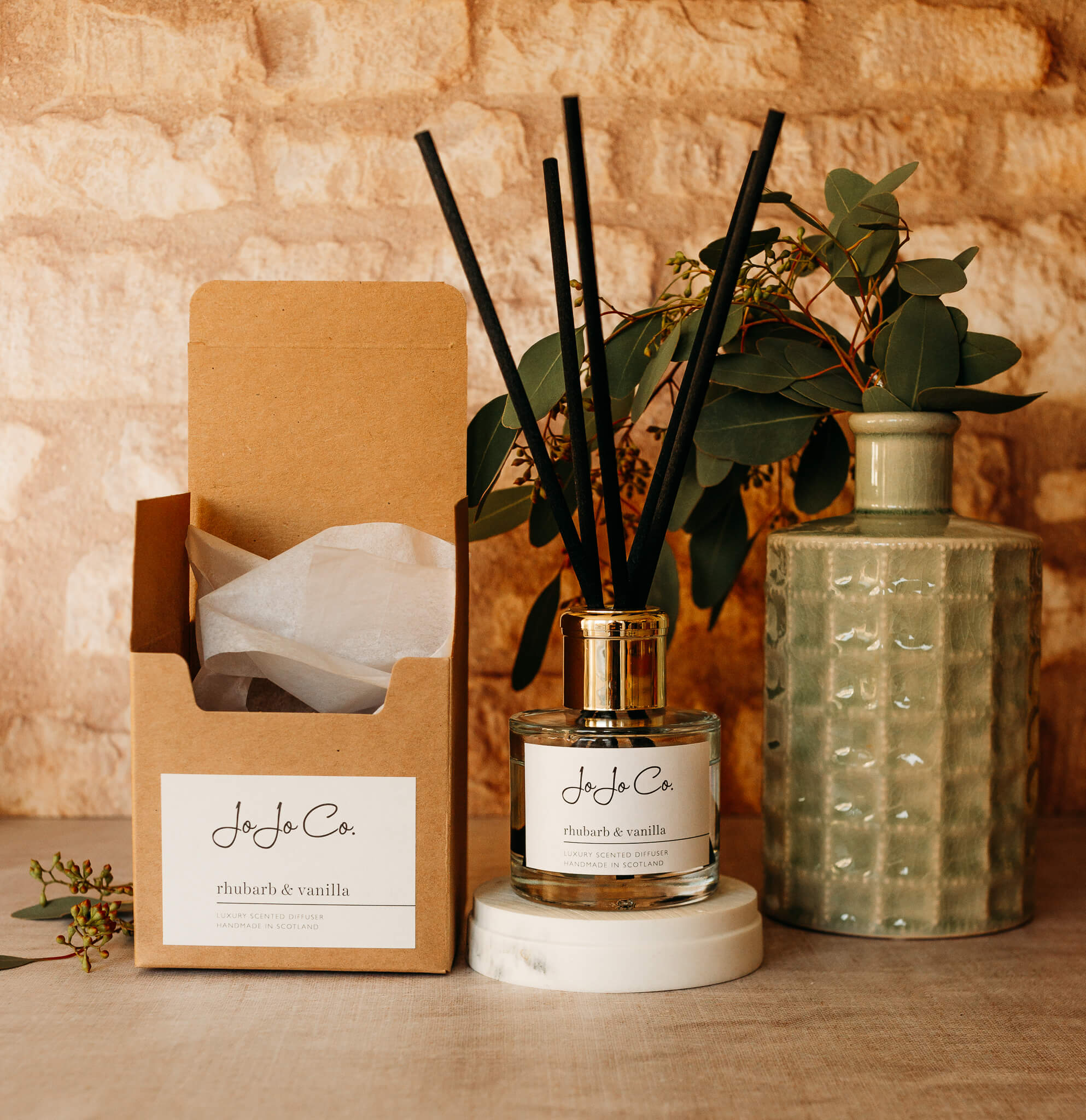 A glass diffuser with cream label, gold lid and black reeds sit on top of a cream candle base. On one side is a green ceramic vase with foliage, on the other side is a cardboard gift box with tissue paper poking out.