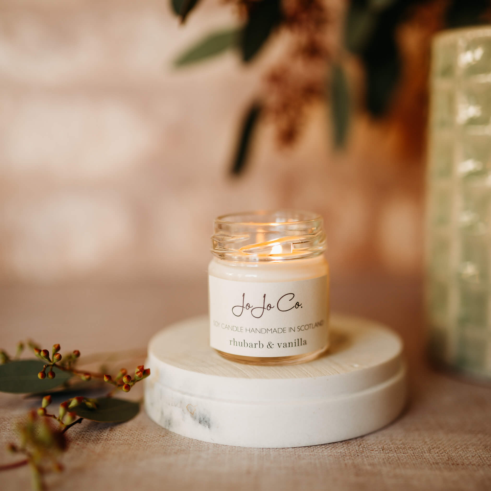 A small, lit candle jar with cream label sits on top of a cream base. In the background is foliage and soft lighting.