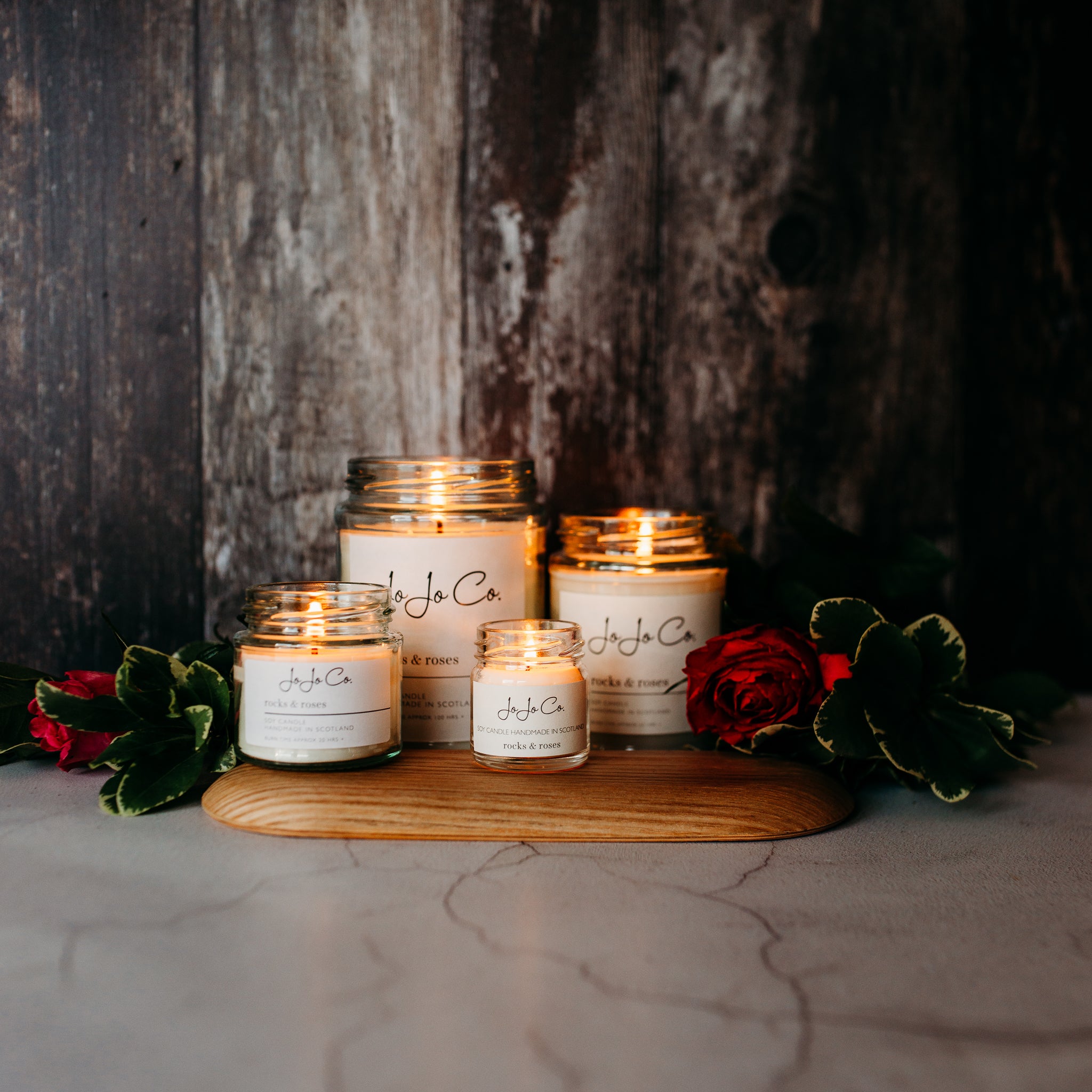 Glass candle jars of various sizes with the label "JoJoCo" on a wooden surface, with flowers and leaves around them creating a moody atmosphere.