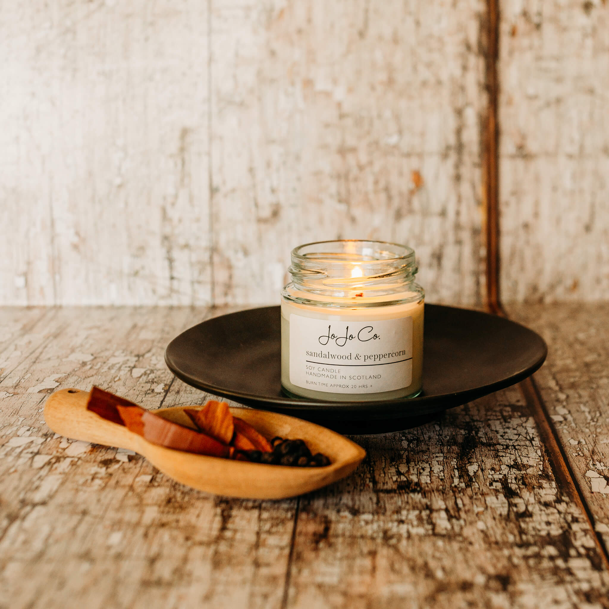 A 20 hour Sandalwood and Peppercorn candle sitting on a black plate with a wooden scoop of sandalwood pieces and peppercorns at the side.