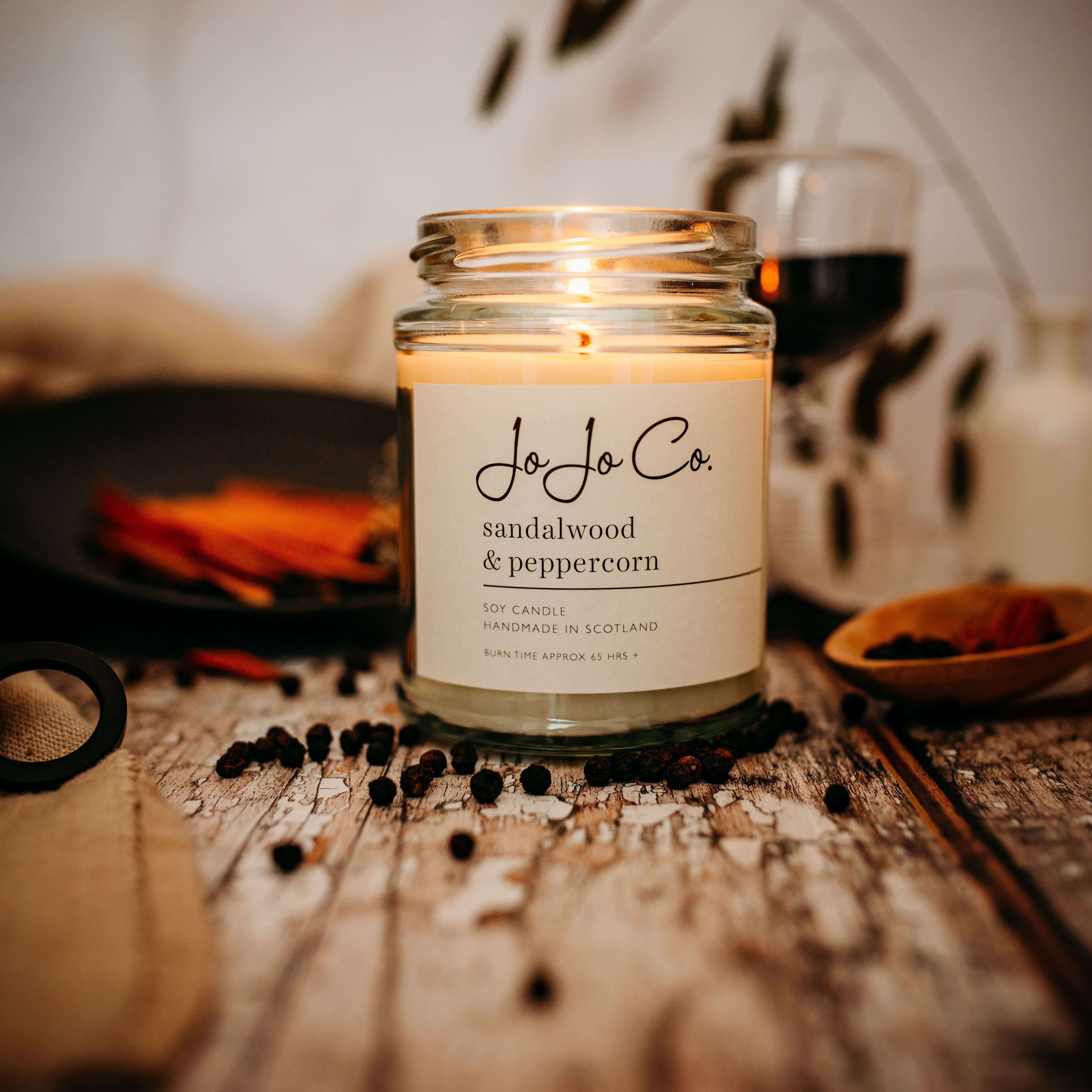 A lit 65 hour Sandalwood and Peppercorn candle in a glass jar with peppercorns scattered in the foreground