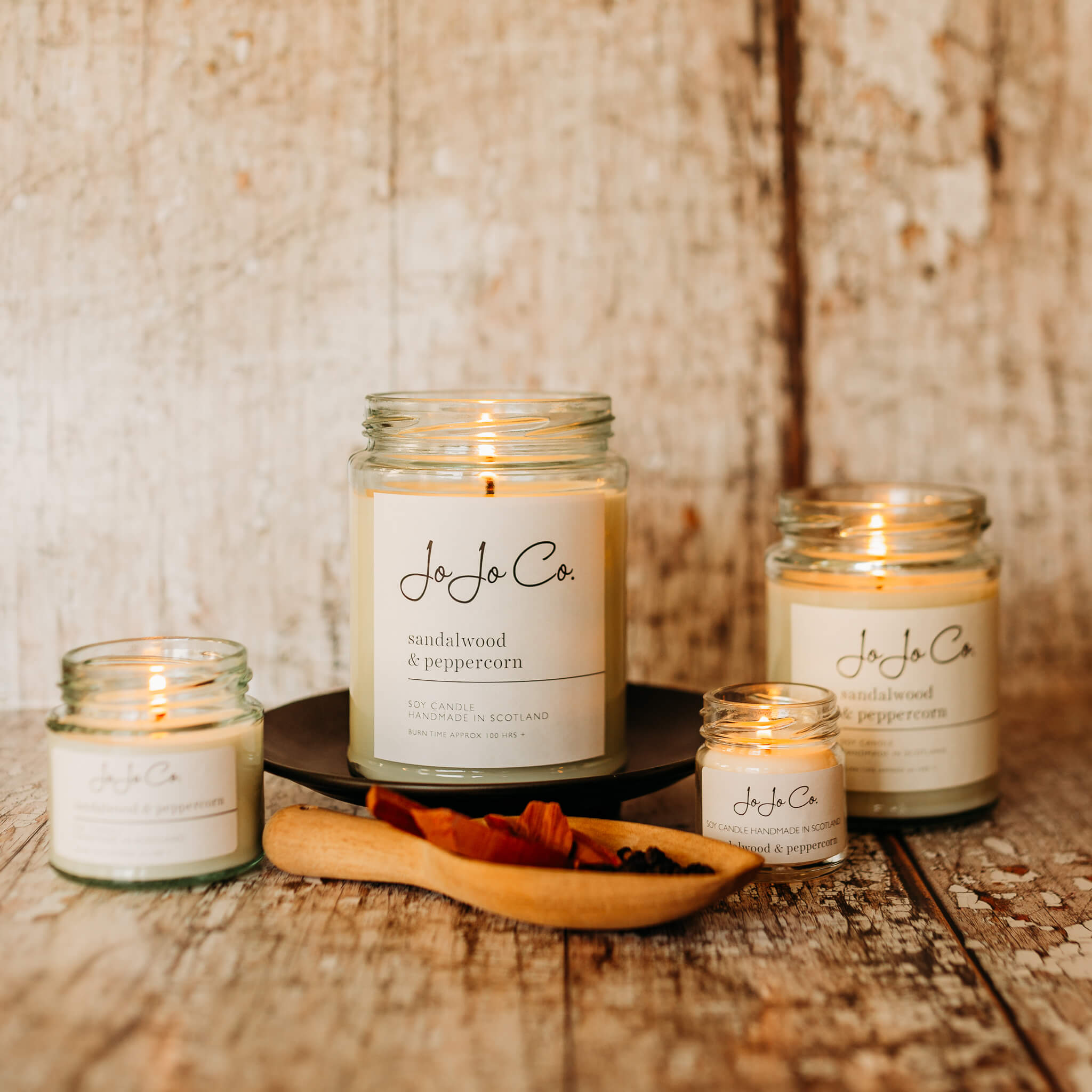Group of JoJo Co Sandalwood and Peppercorn candles in glass jars in a variety of sizes on a wooden textured backdrop
