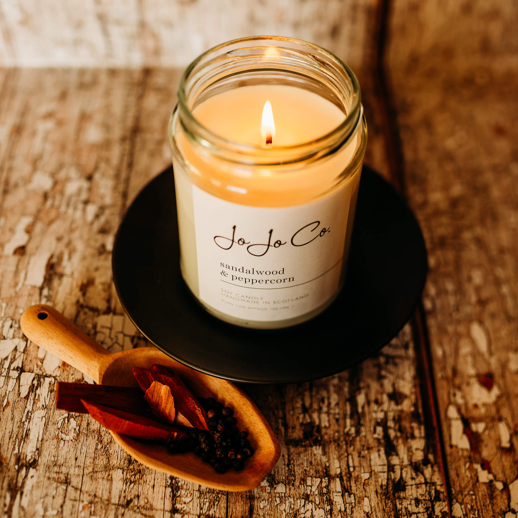 Looking down on a 100 hour Sandalwood and Peppercorn candle in a glass jar with a wooden scoop with some pieces of sandalwood and peppercorns