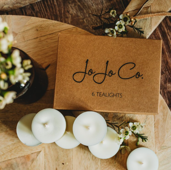 A small cardboard gift box with JoJo Co. branding sits on a table surrounded by white flowers and unlit white tealight candles.
