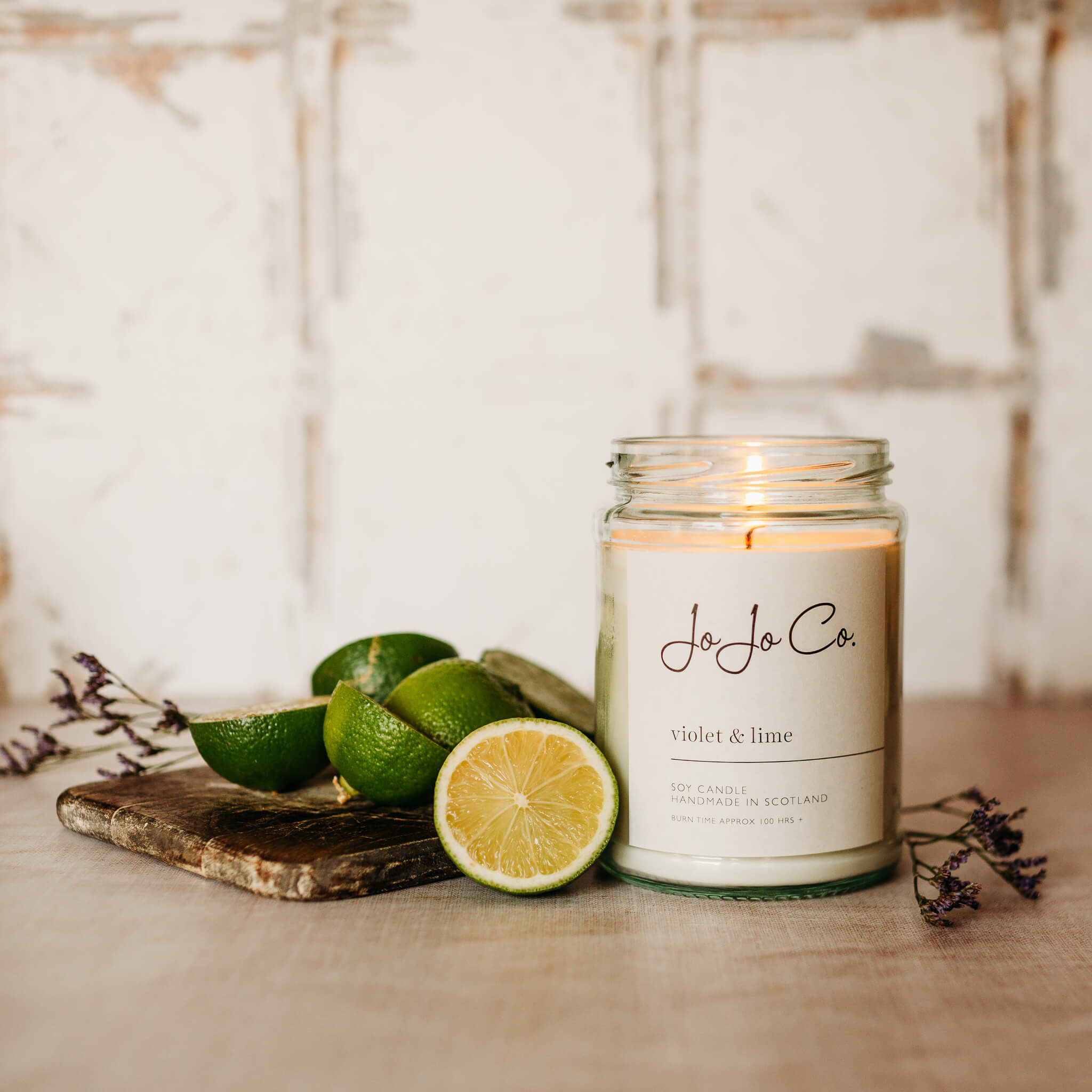 A large JoJo Co. Violet & Lime candle in a glass jar and white label sits in front of a white background. Slices of lime and dried flowers surround the candle.