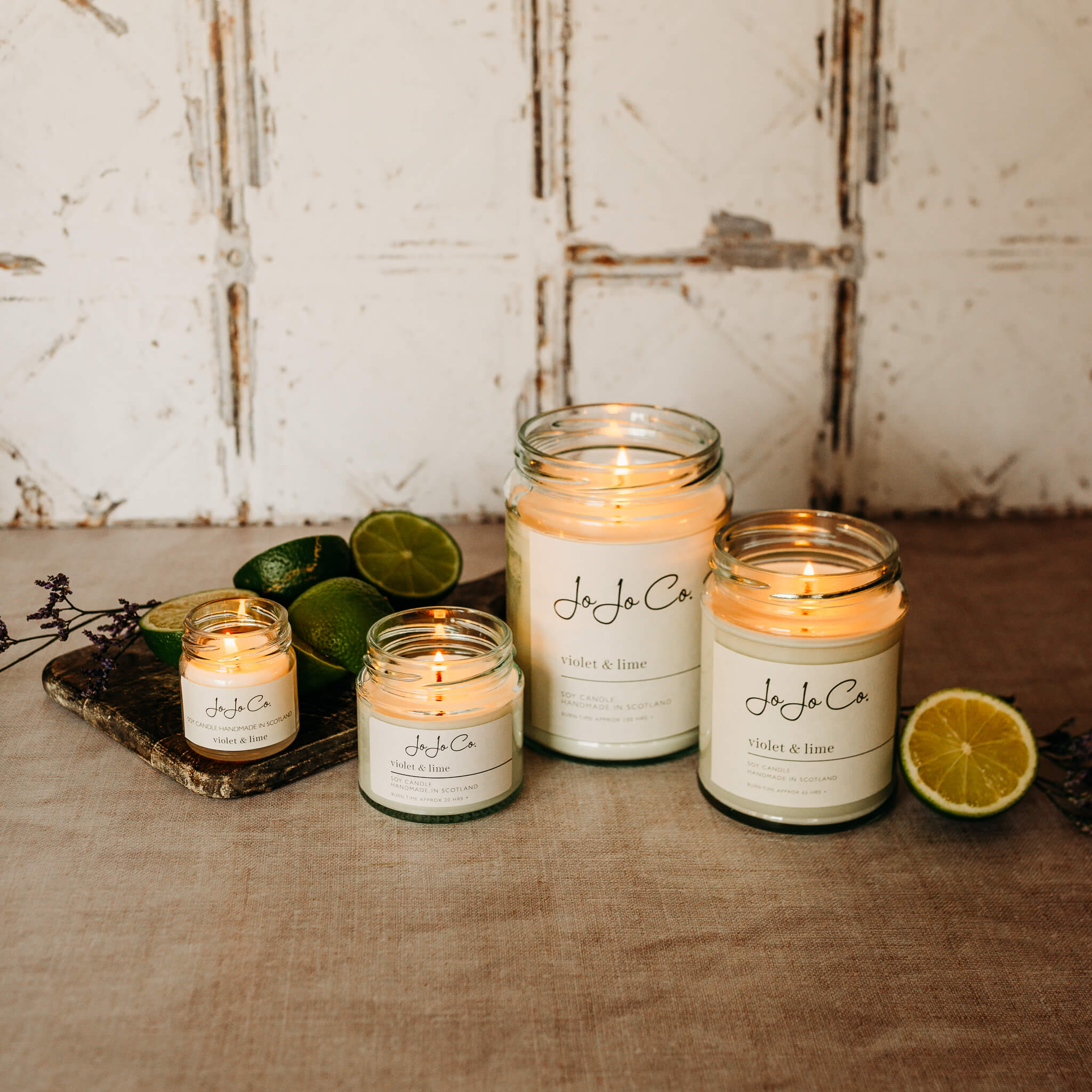 A selection of JoJo Co. Violet & Lime candles sits on a wooden floor with white tiles in the background. Slices of lime and dried flowers surround the glass jar candles.
