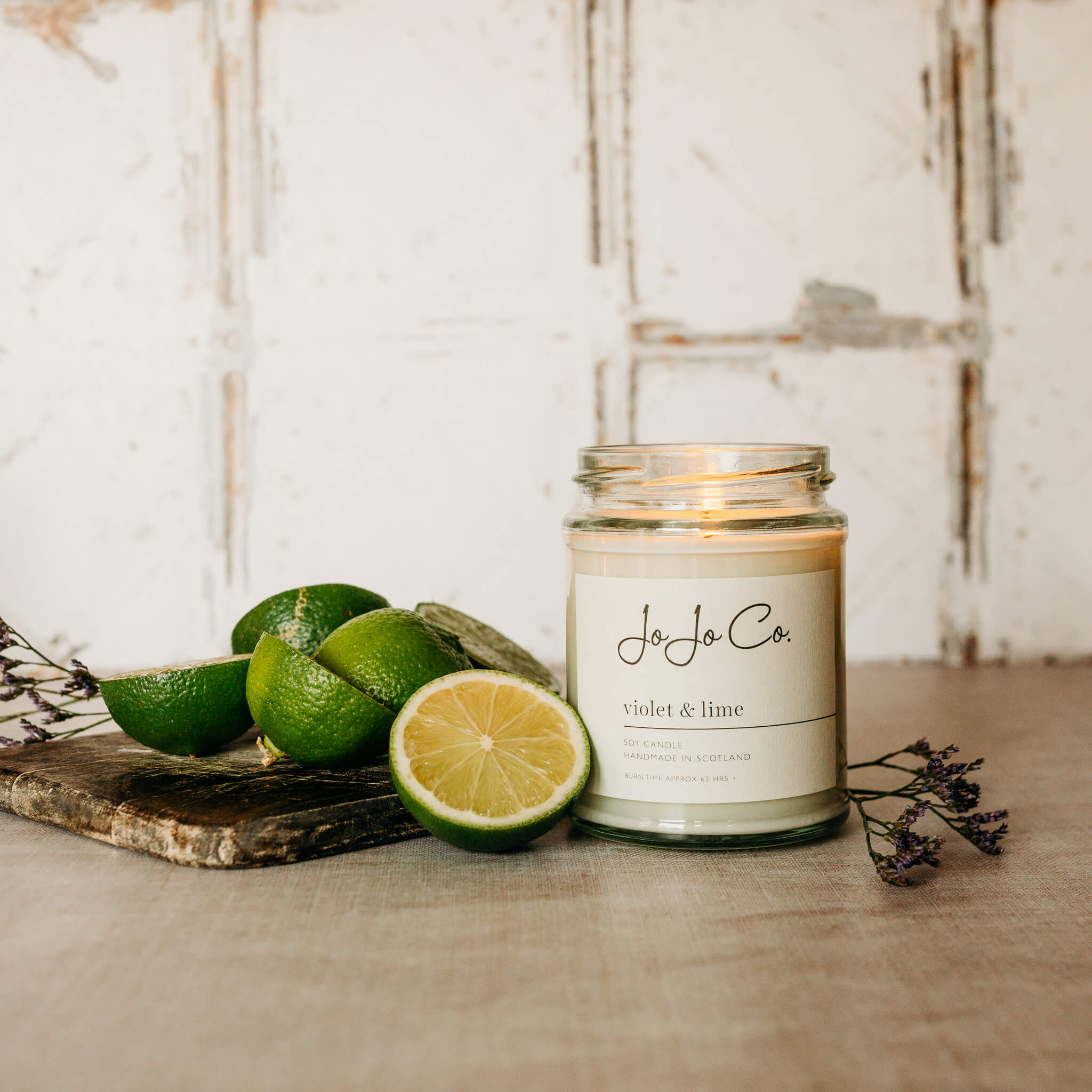 A large white labelled JoJo Co. Violet & Lime candle sits on a beige surface with white tiled background. A wooden board, dried leaves and slices of lime surround it.