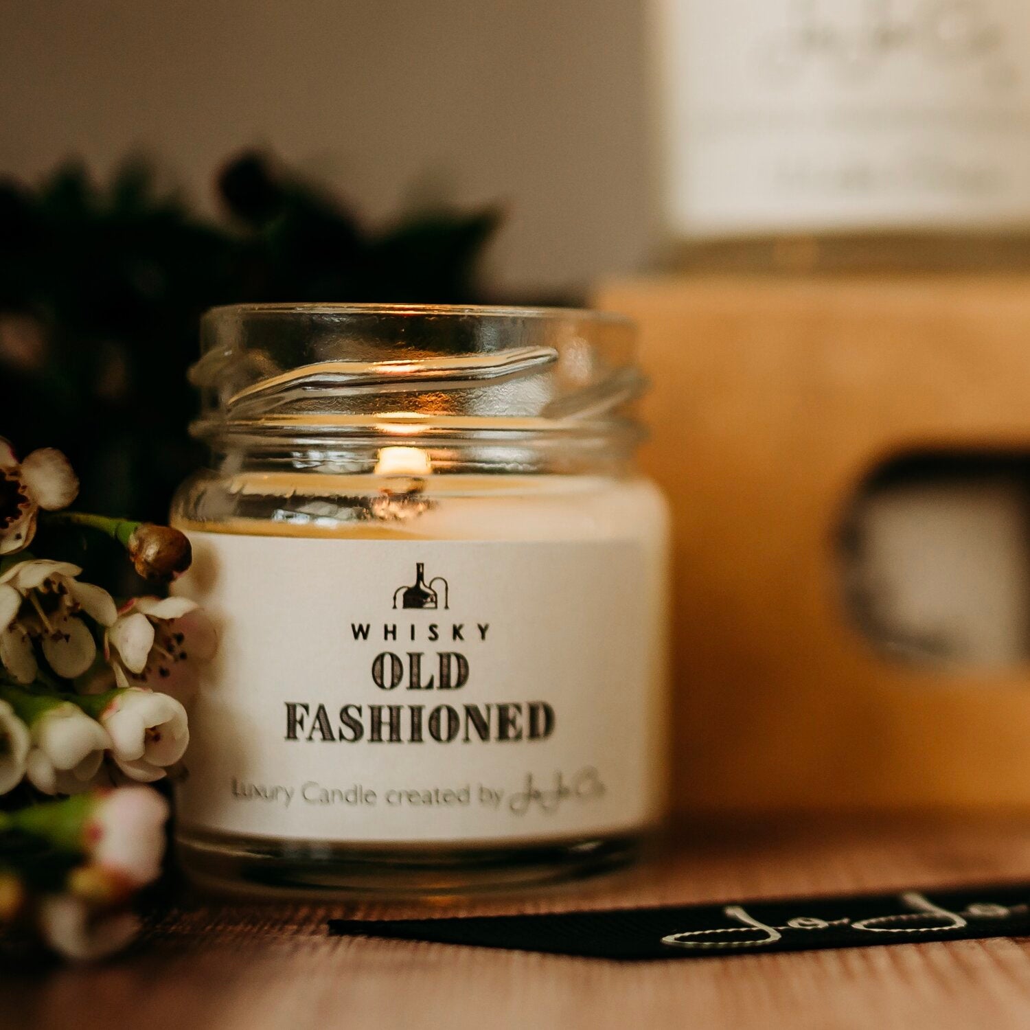 A mini JoJo Co. Whisky Old Fashioned glass jar candle sits beside white flowers.