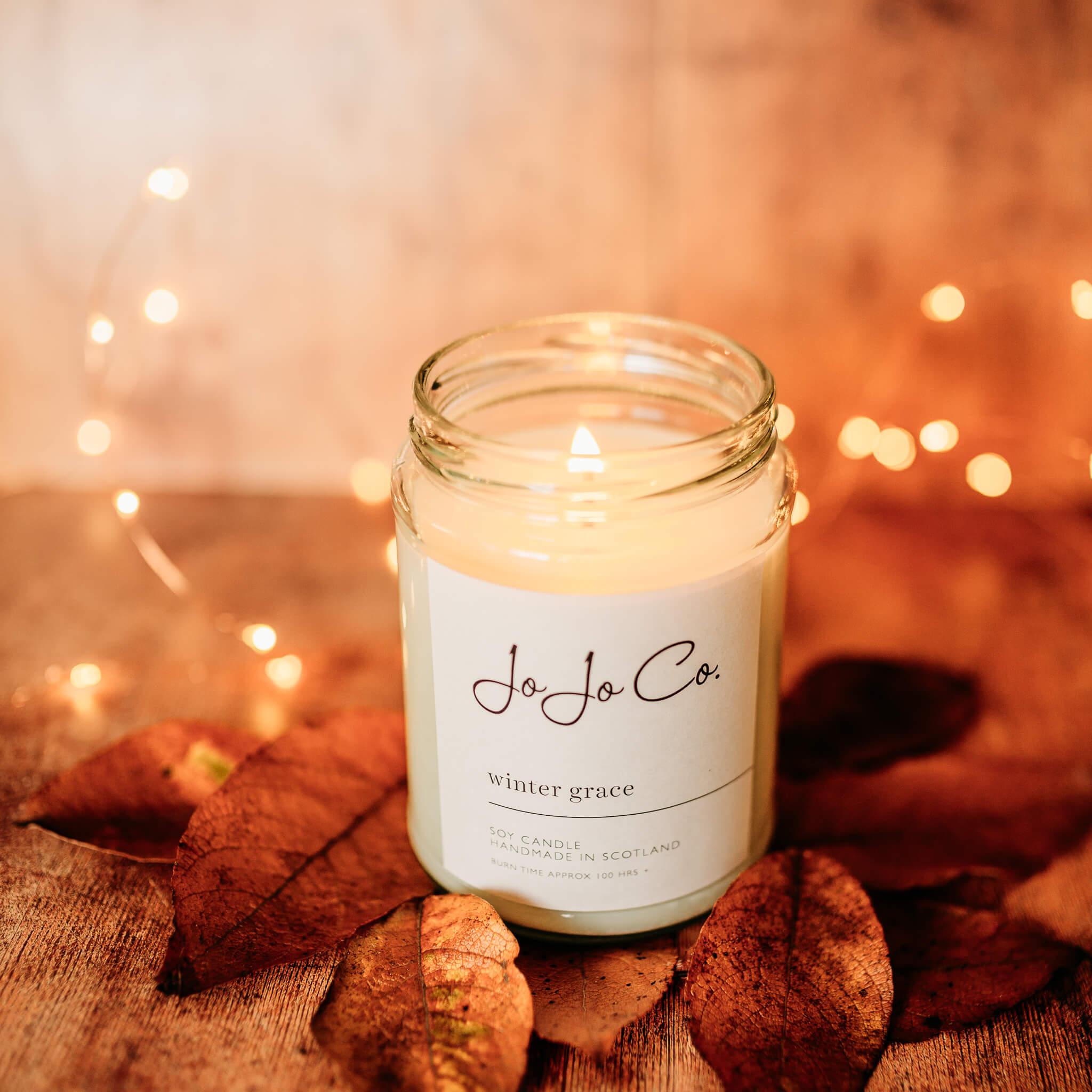 A large 100 hour Winter Grace JoJo Co candle with white label and flickering flame sits in front of twinkling fairy lights, surrounded by dried leaves.