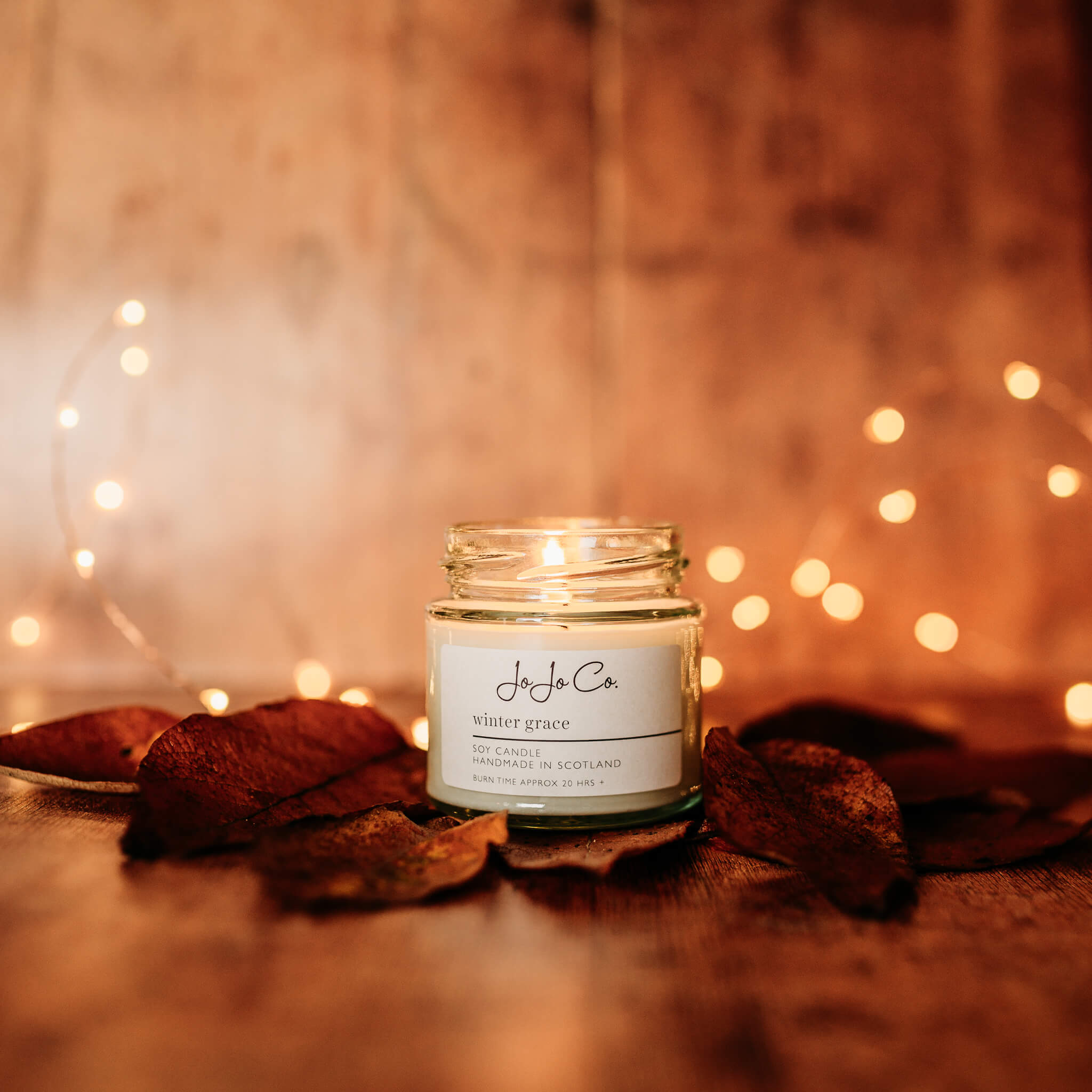 A 20 hour Winter Grace JoJo Co. candle sits beside fallen leaves and sparkling fairy lights. The light from the flame casts a warm glow.