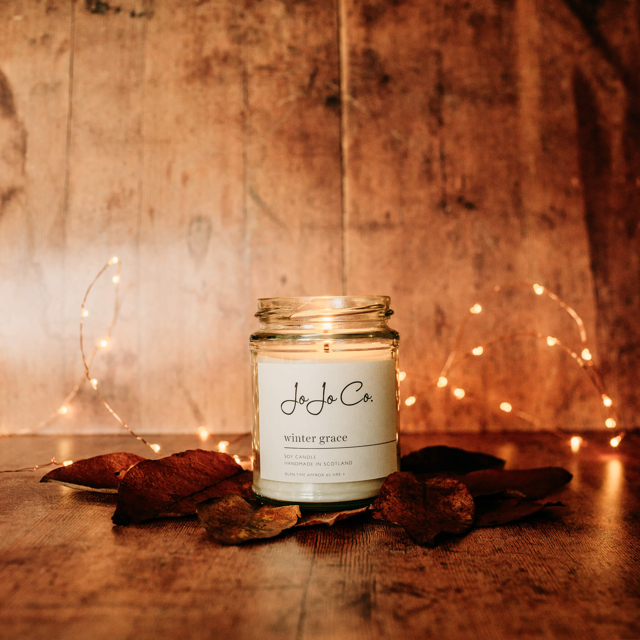 A large JoJo Co Winter Grace candle sits in front of sparkling fairy lights surrounded by leaves on a warm wooden floor.