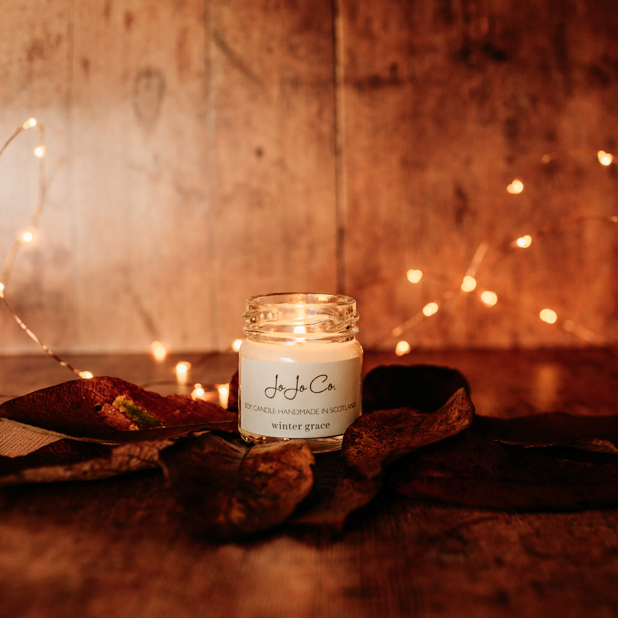 A mini glass JoJo Co. candle with flickering flames. Surrounded by dried leaves and twinkling fairy lights.