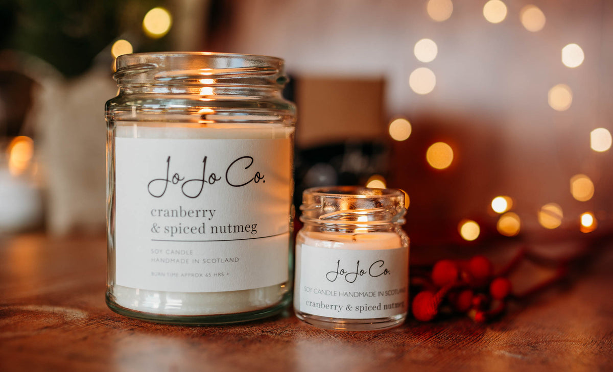 Two JoJo Co. scented candles in glass jars, one large and one small, displayed on a wooden surface with warm bokeh string lights in the background.